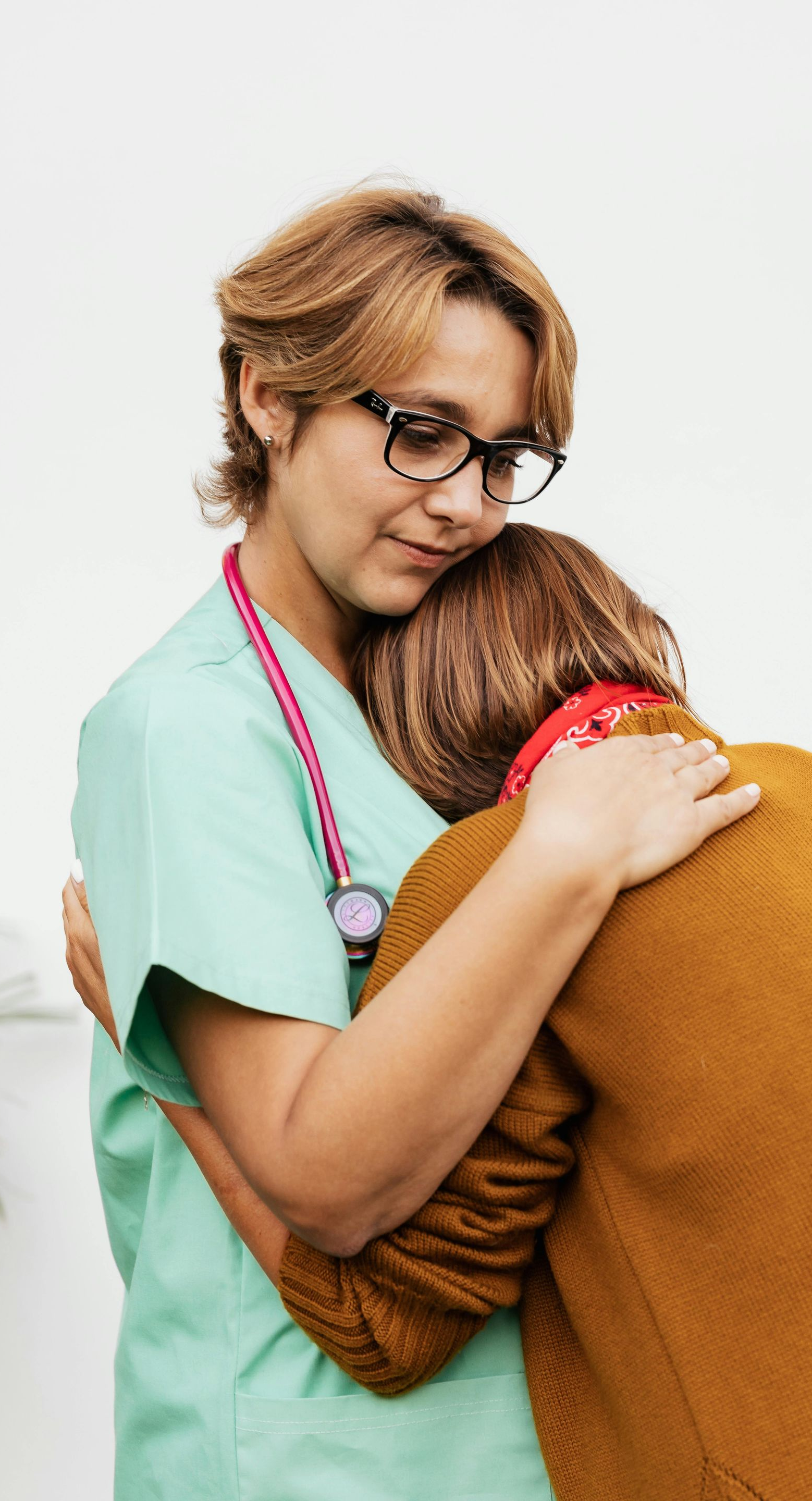 Nurse in scrubs hugs a person wearing a brown sweater; both have compassionate expressions.