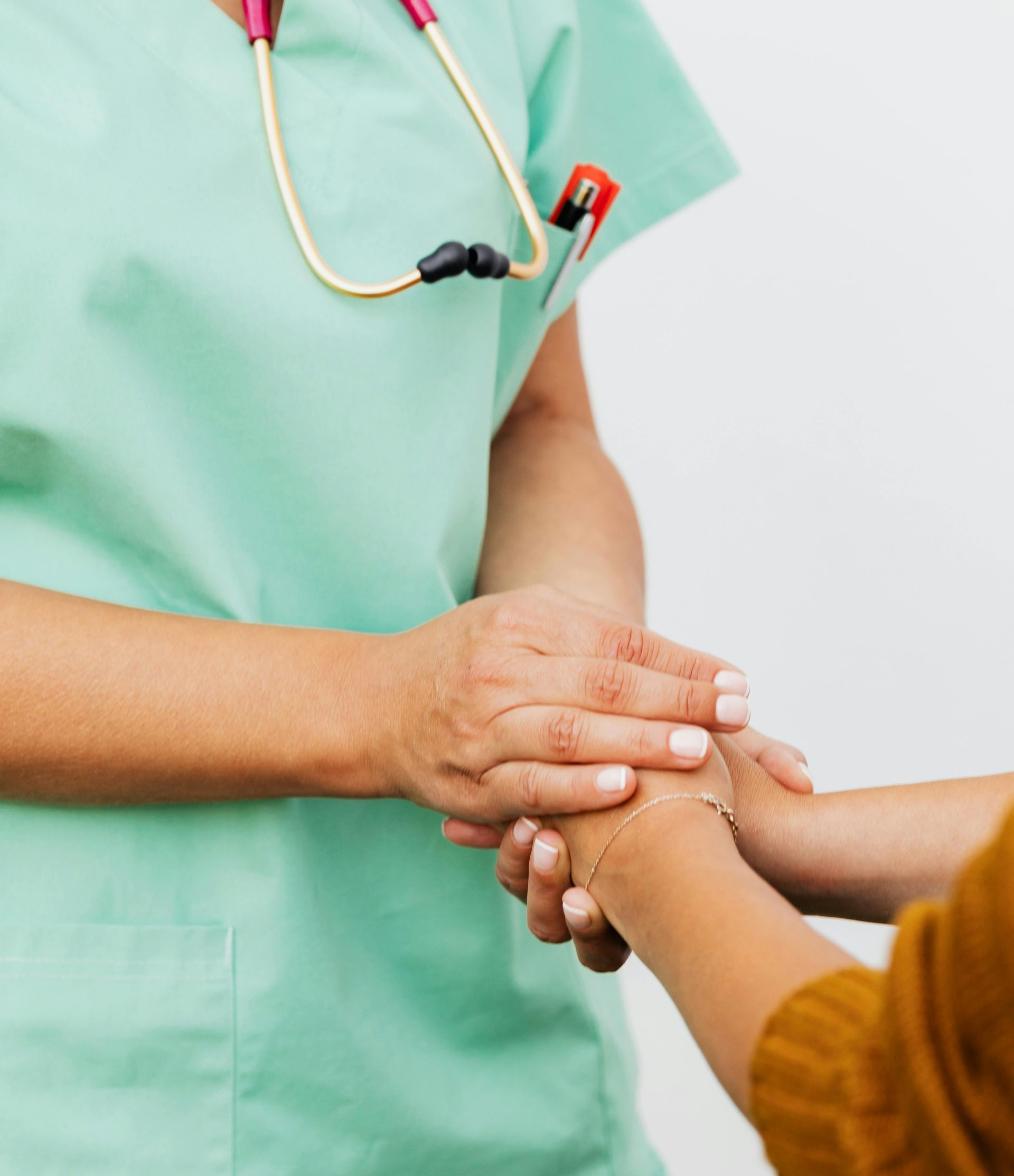 Nurse in green scrubs comforts a patient, holding their hand. Pink stethoscope rests on the nurse's chest.