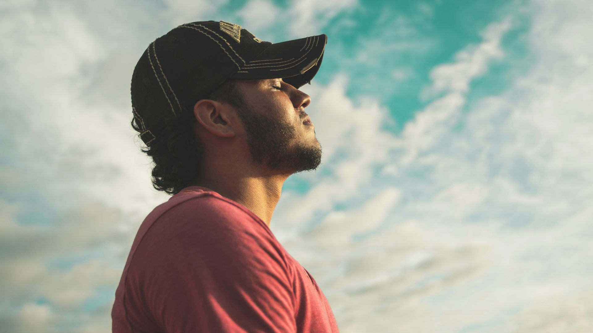 Man in red shirt and cap looking up at a cloudy sky.