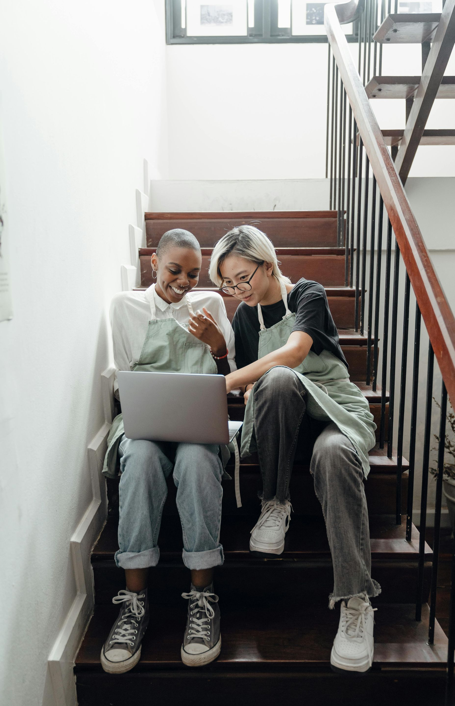 Two people wearing aprons sitting on wooden stairs, looking at a laptop and smiling.