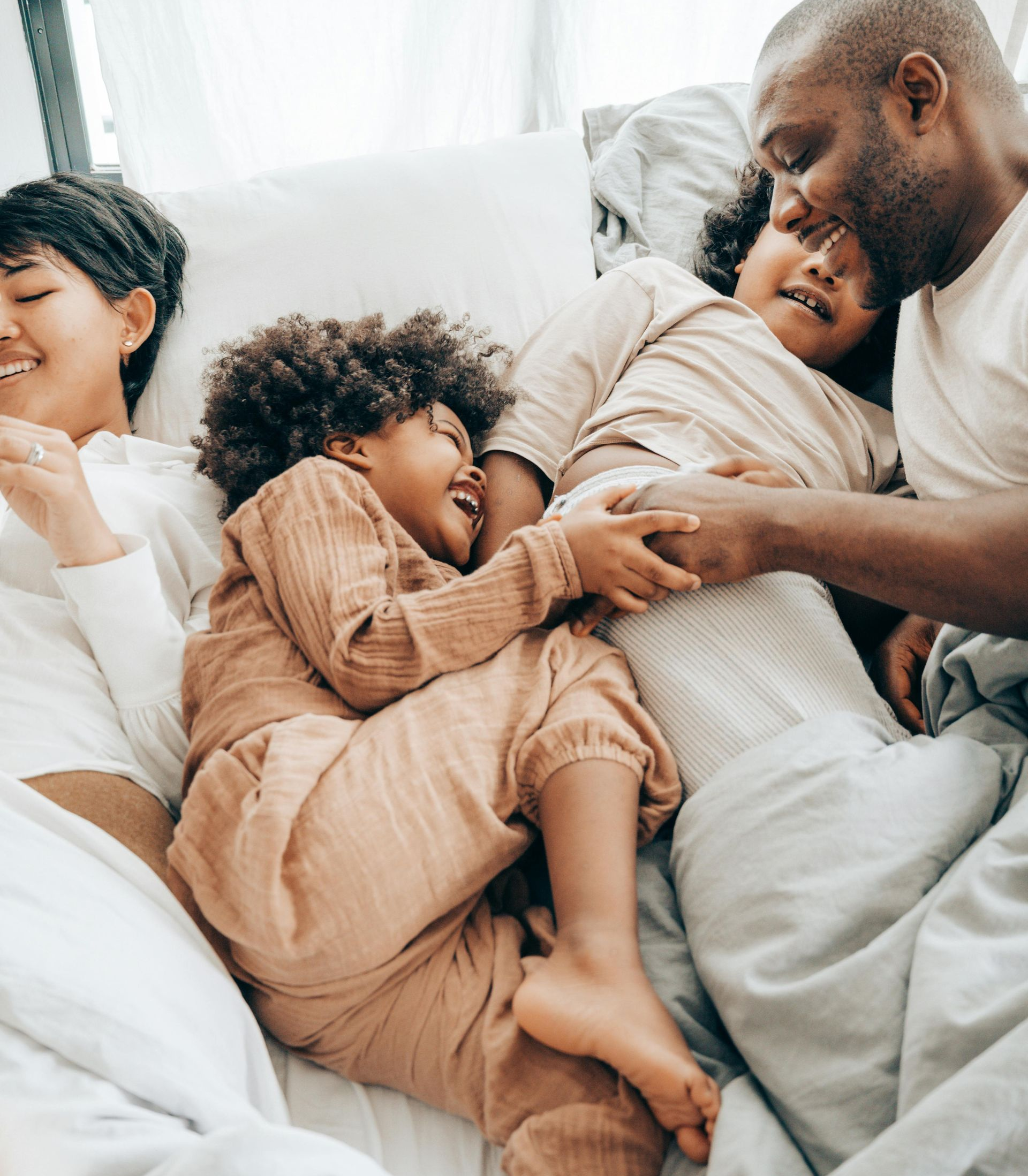 Family of four laughing together on a bed with light-colored bedding.