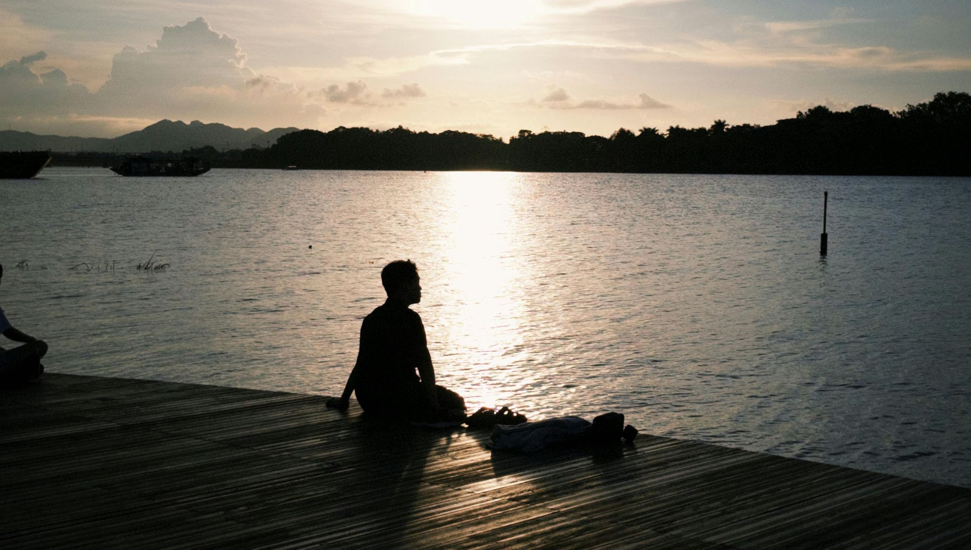 Person silhouetted on a dock, gazing at a shimmering sunset over water, with boats and trees in the background.
