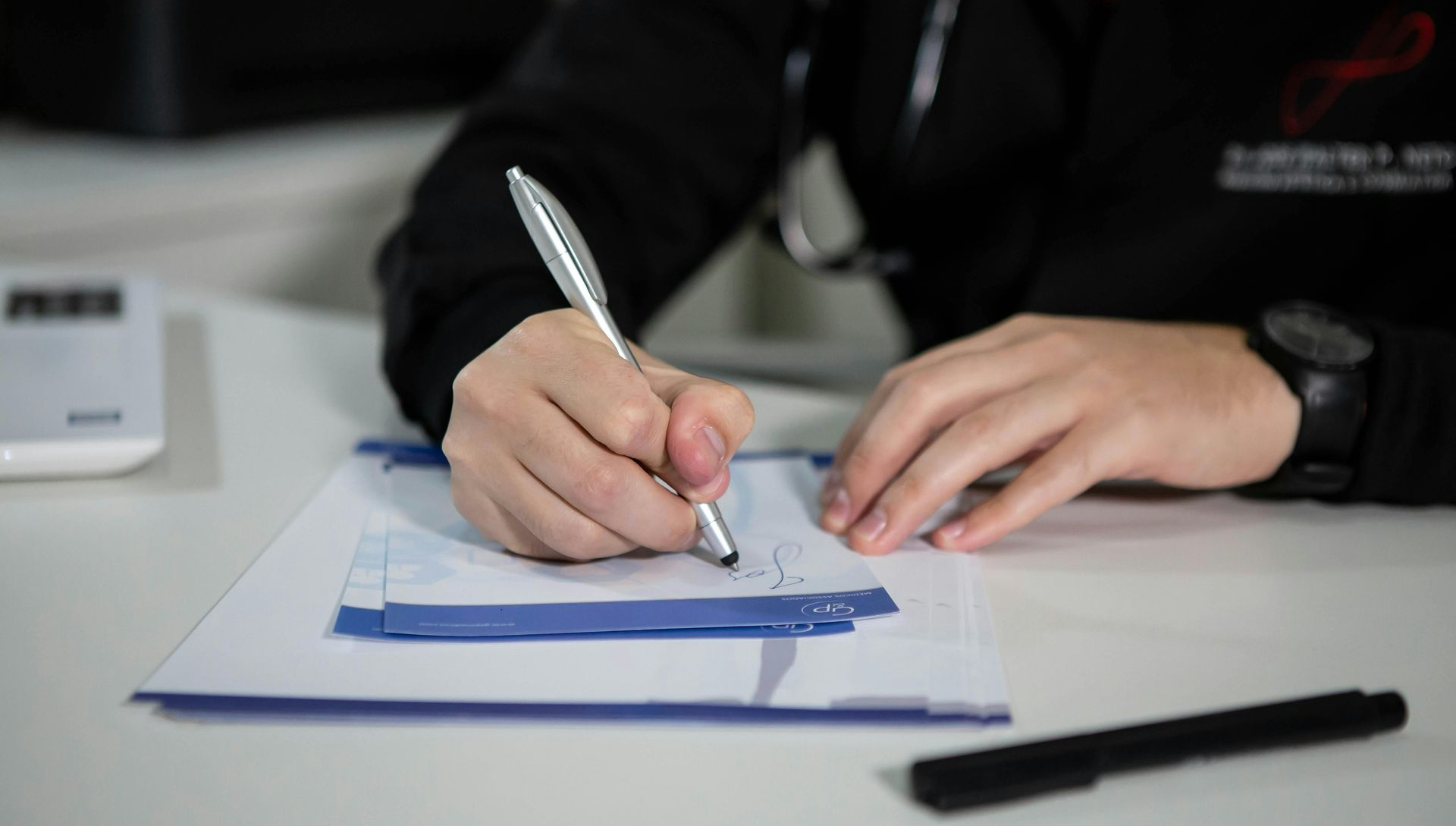 Doctor writing on a clipboard with a pen on a white desk; stethoscope visible.