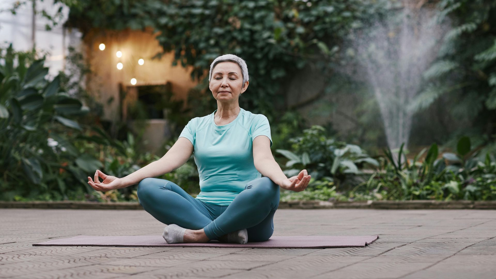 Woman meditating on a mat outdoors, eyes closed, hands in Gyan Mudra. Green foliage and fountain in the background.
