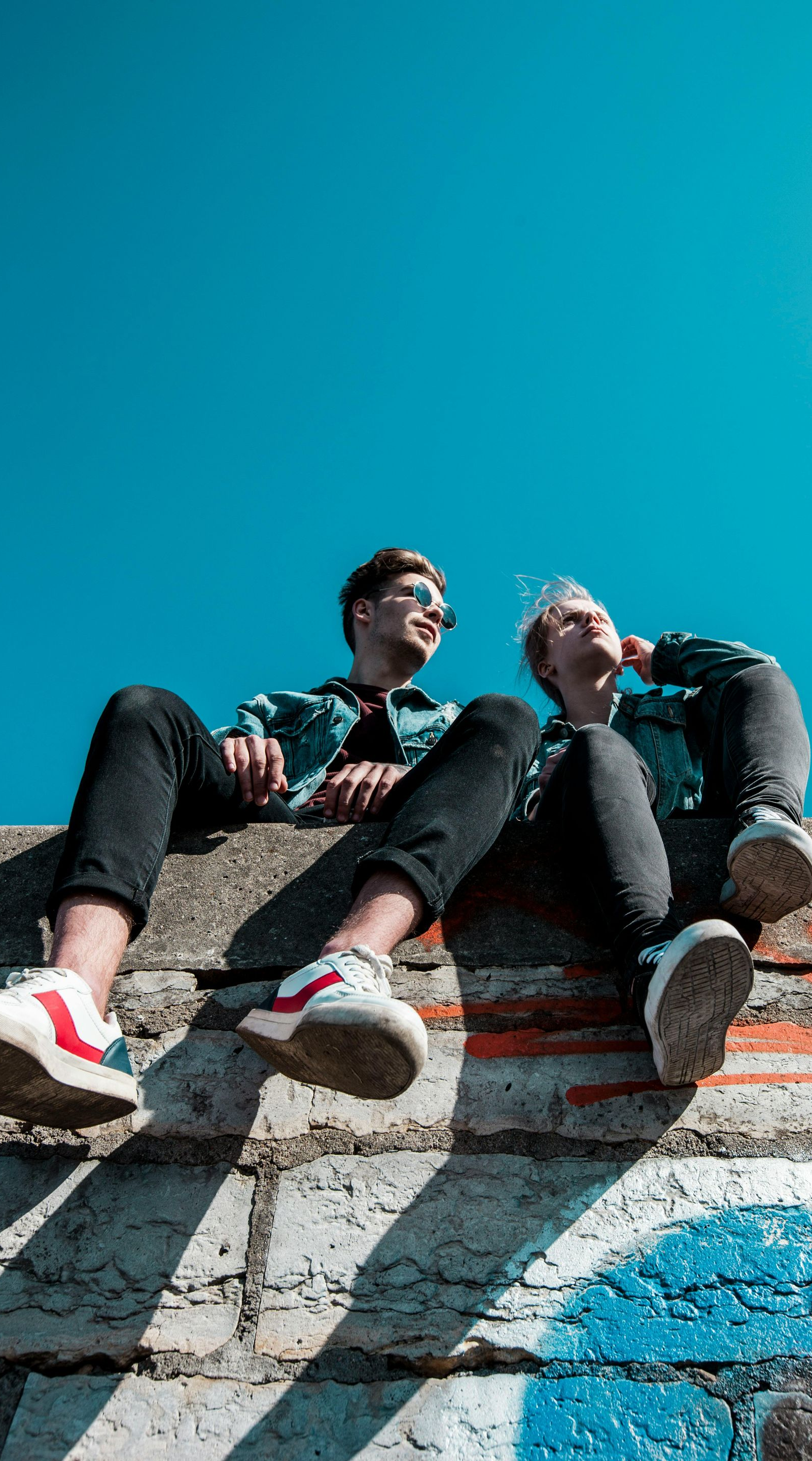 Two people in denim jackets and jeans sit on a brick wall against a blue sky.