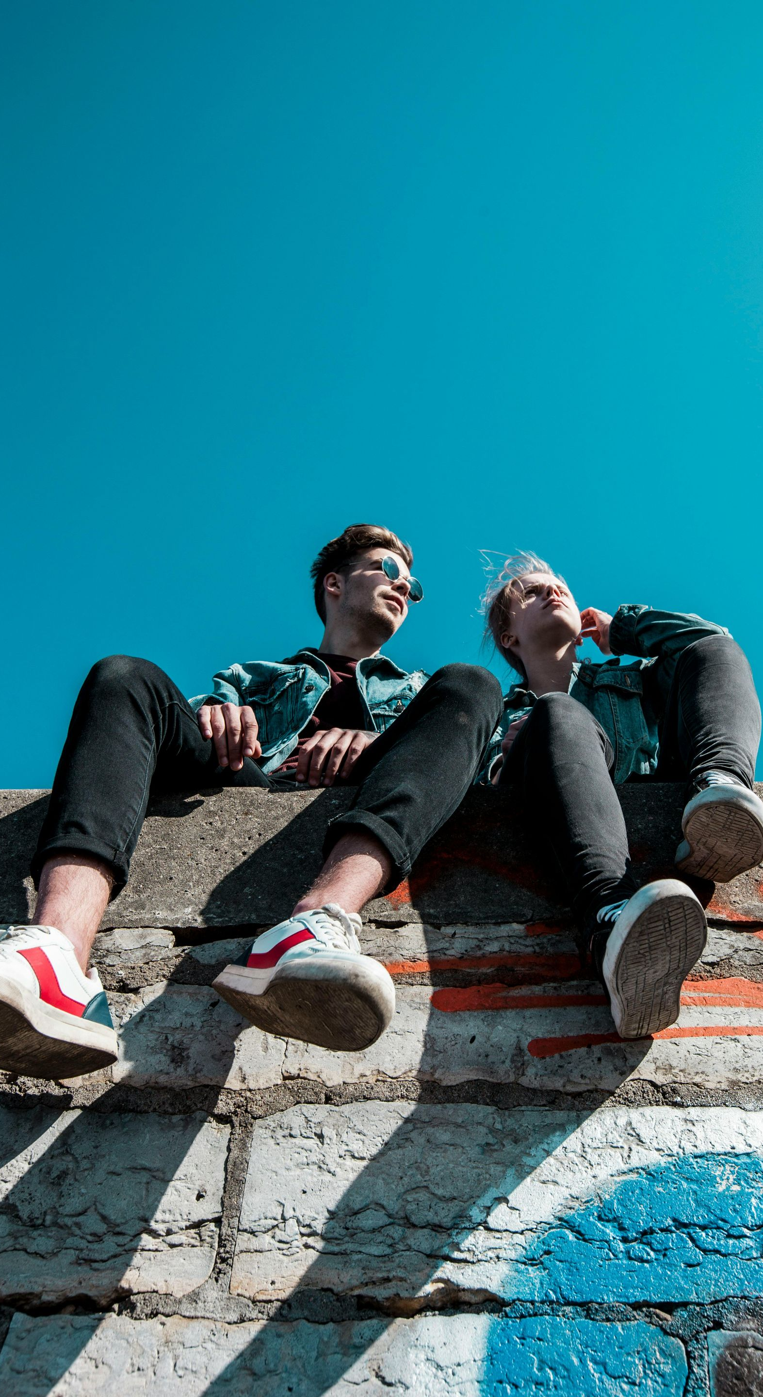 Two people in denim jackets and jeans sit on a brick wall against a blue sky.