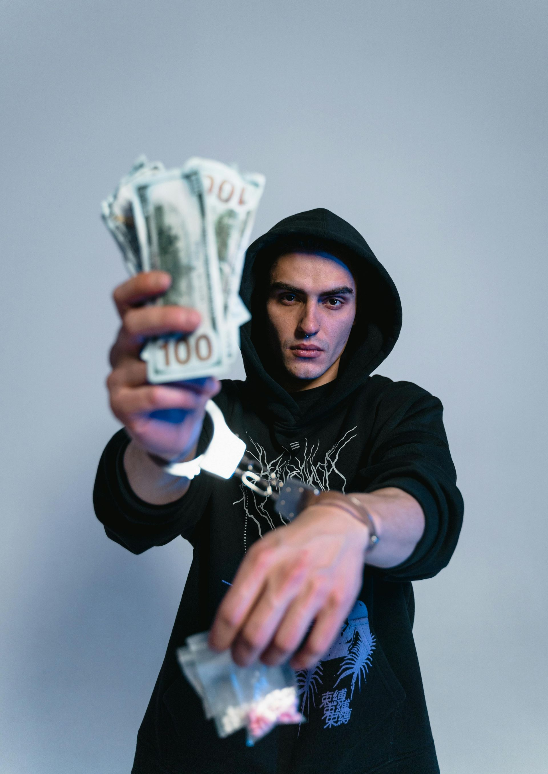 Man in hoodie holds money and bag with white powder, wearing handcuffs.