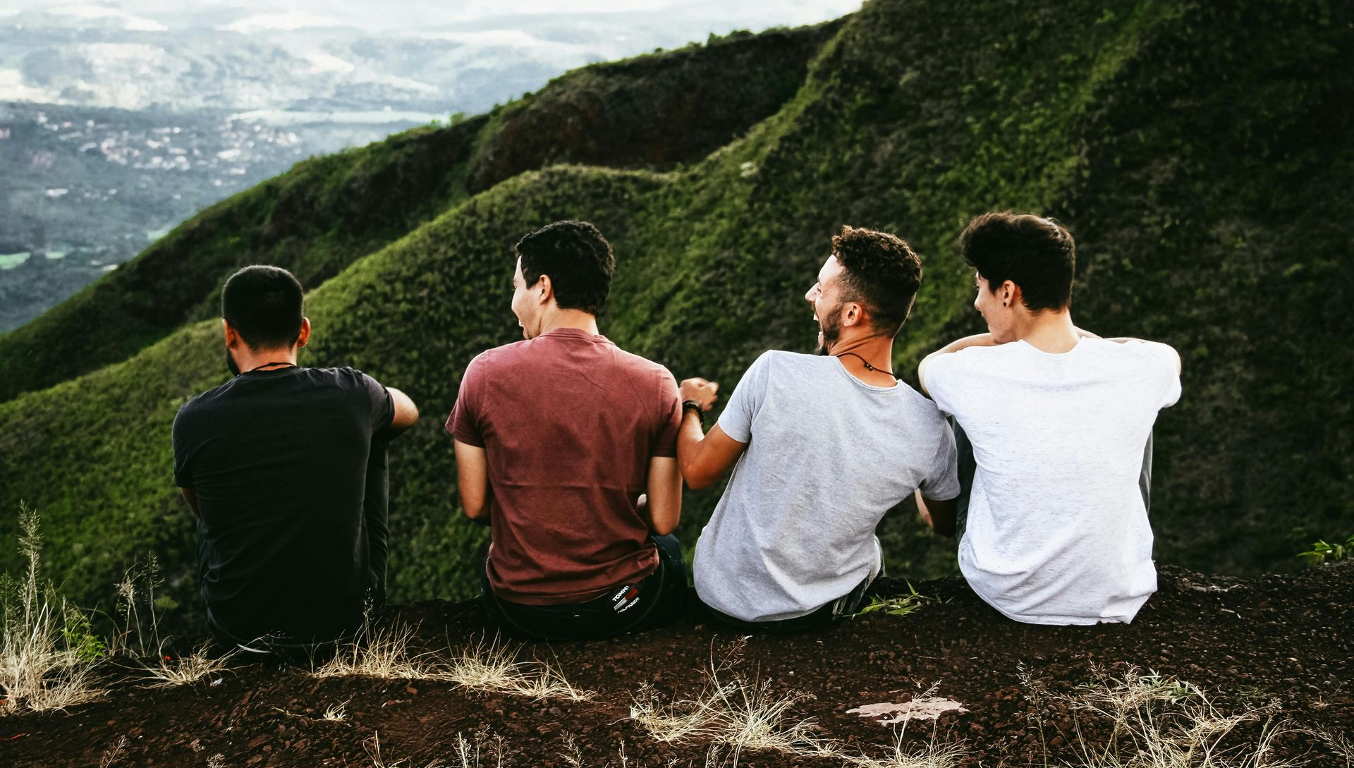 Four people seated on a hillside, overlooking a landscape. Their backs are turned, arms around each other.