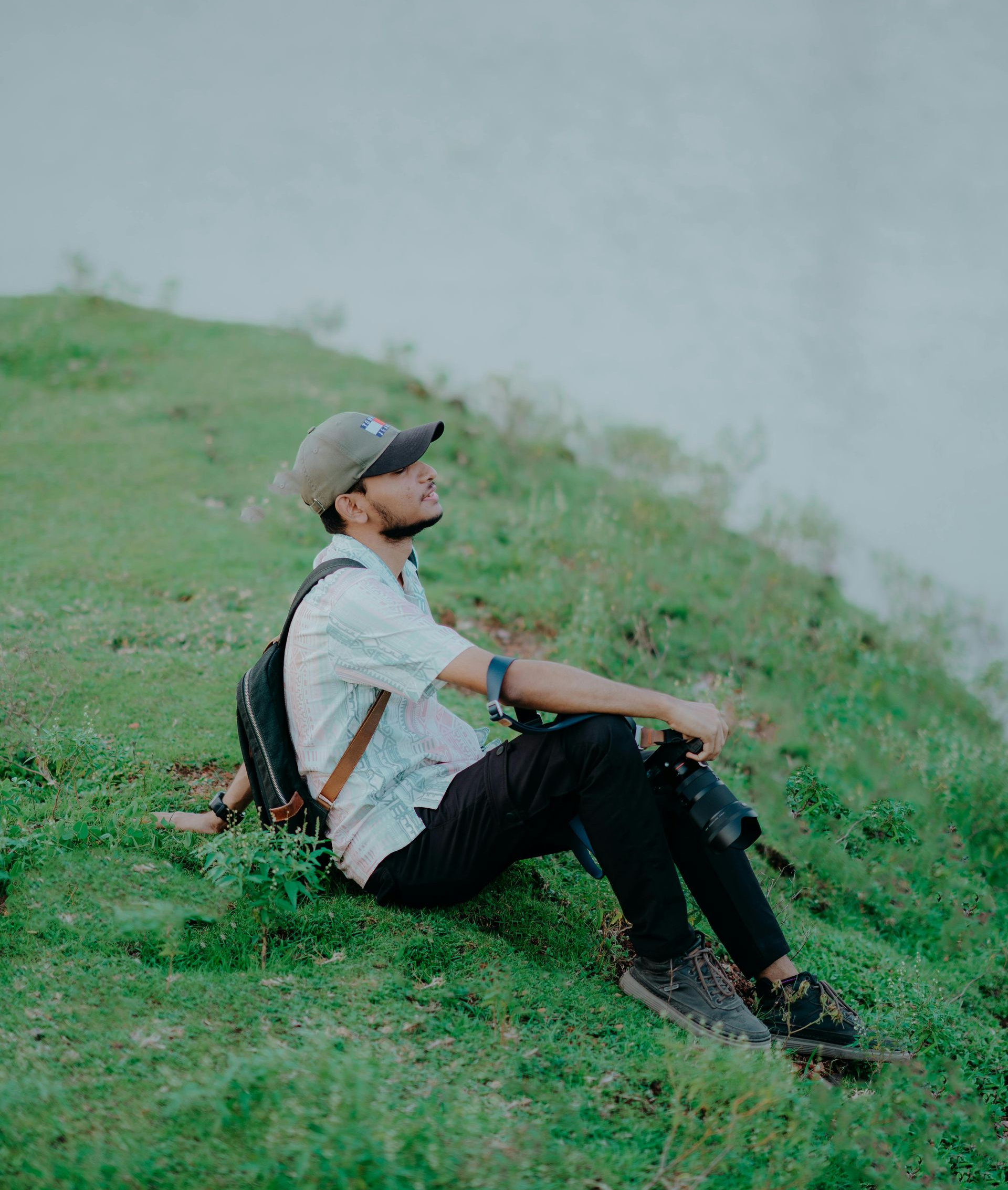 Man with backpack sits on grassy hill, looking up. Holds camera, wearing cap, white shirt, dark pants.