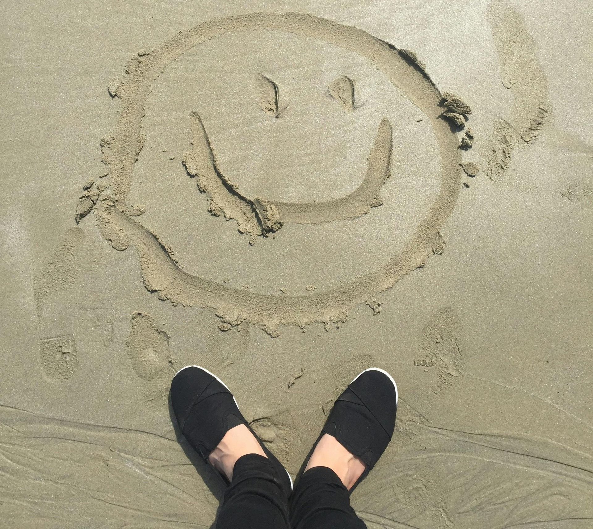 Person's feet in black shoes stand over a smiley face drawn in sand.