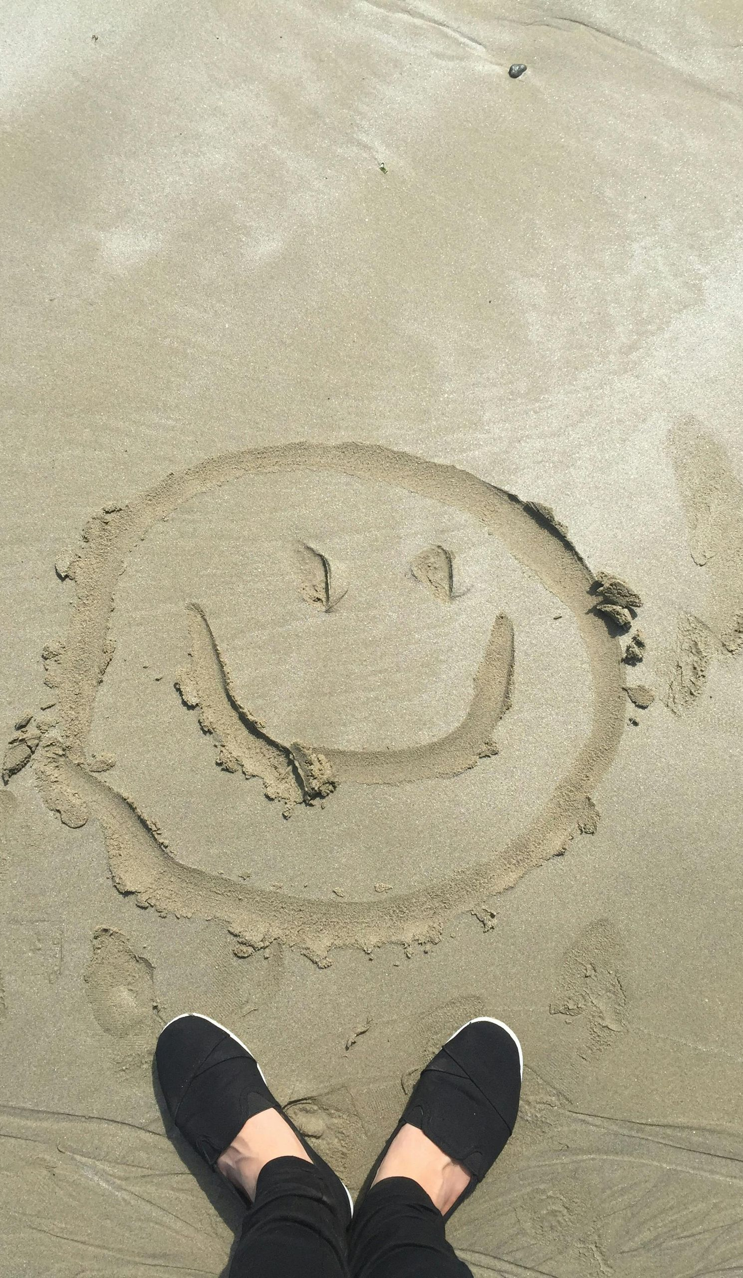 Smiley face drawn in sand; person wearing black shoes stands near it at the beach.
