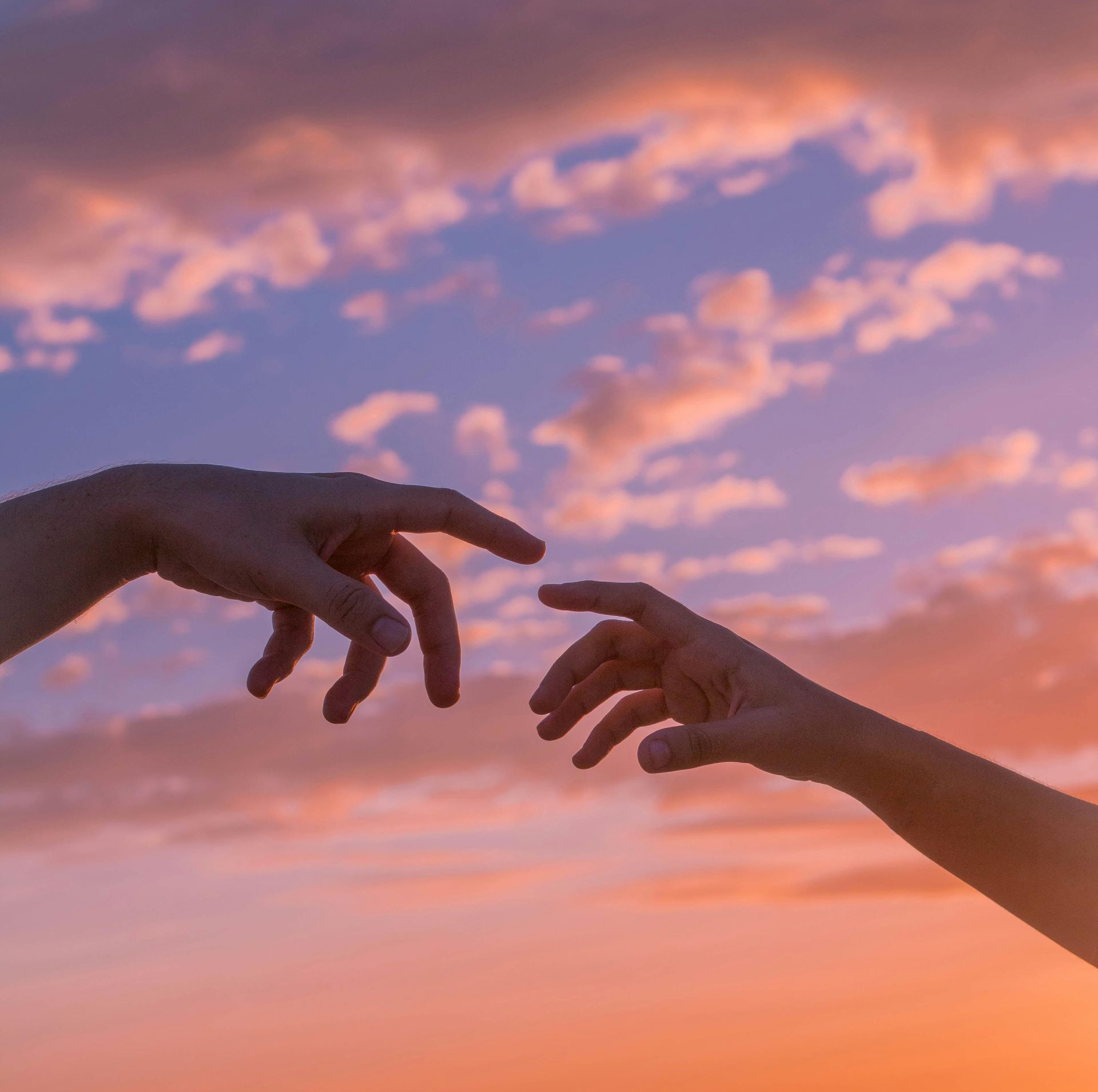 Two hands reaching towards each other, nearly touching, against a sunset sky of pinks, oranges, and blues.