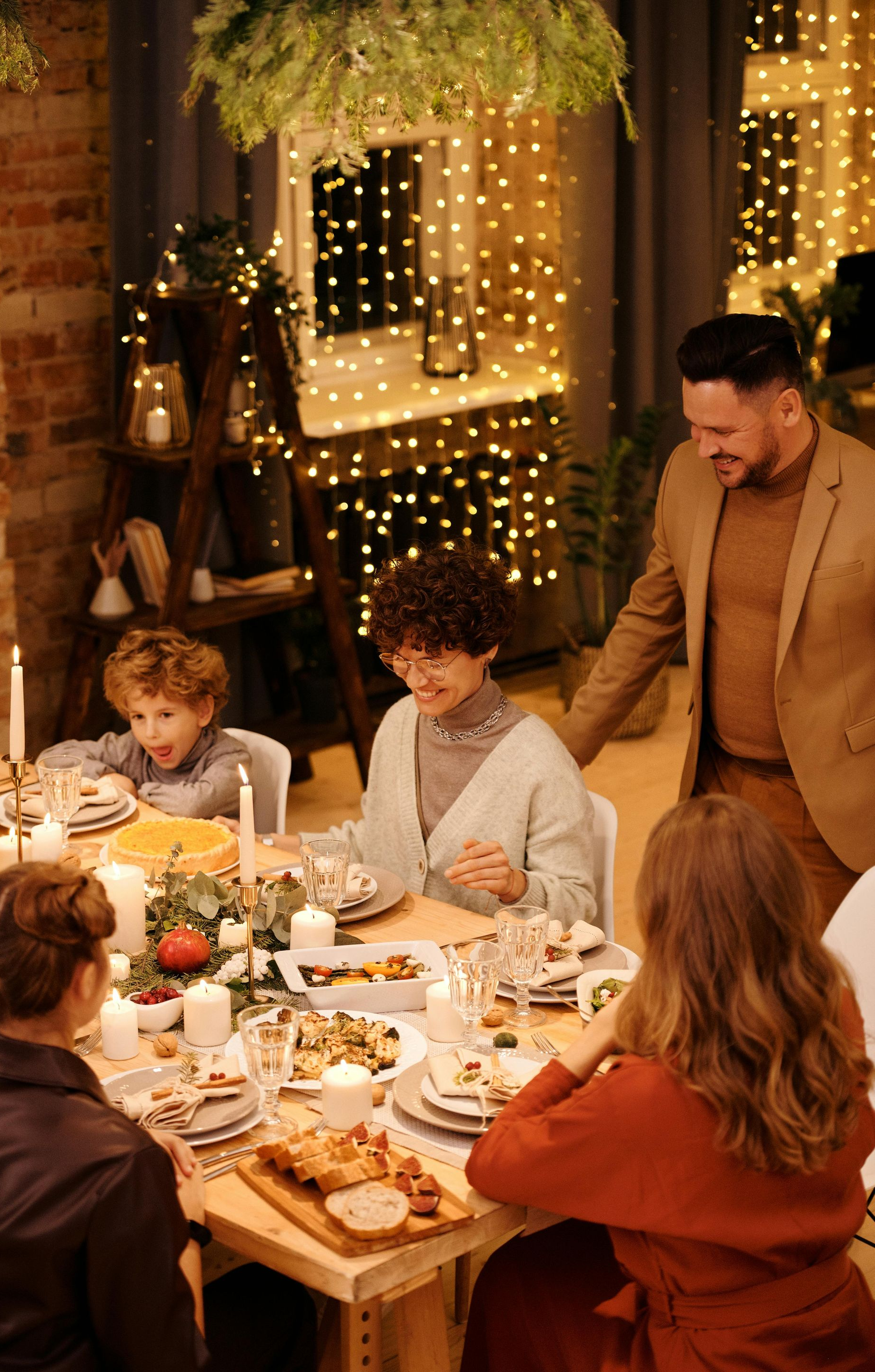Family gathered around a festively decorated table, illuminated by lights. Some are smiling, and one man stands nearby.