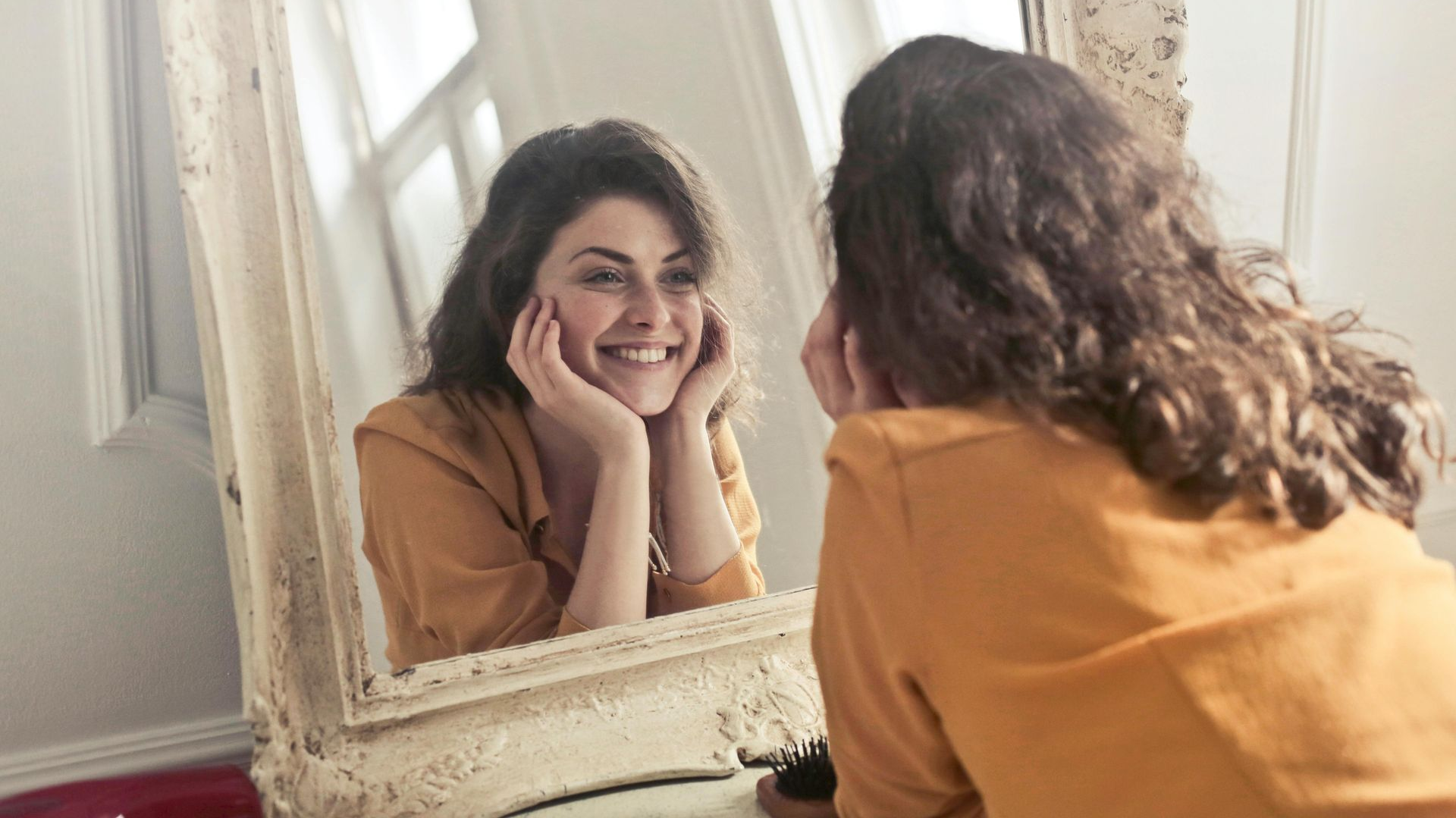 Woman in yellow shirt smiles at herself in ornate, white framed mirror.