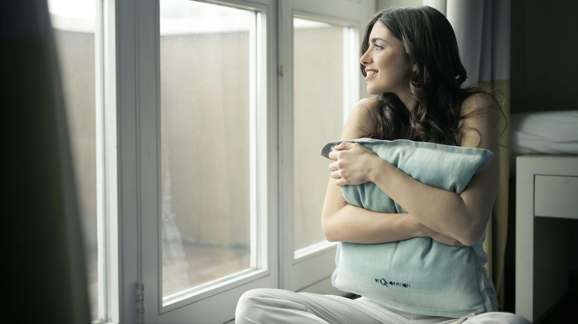 Woman holding a blue pillow, sitting by a window, looking outside and smiling.