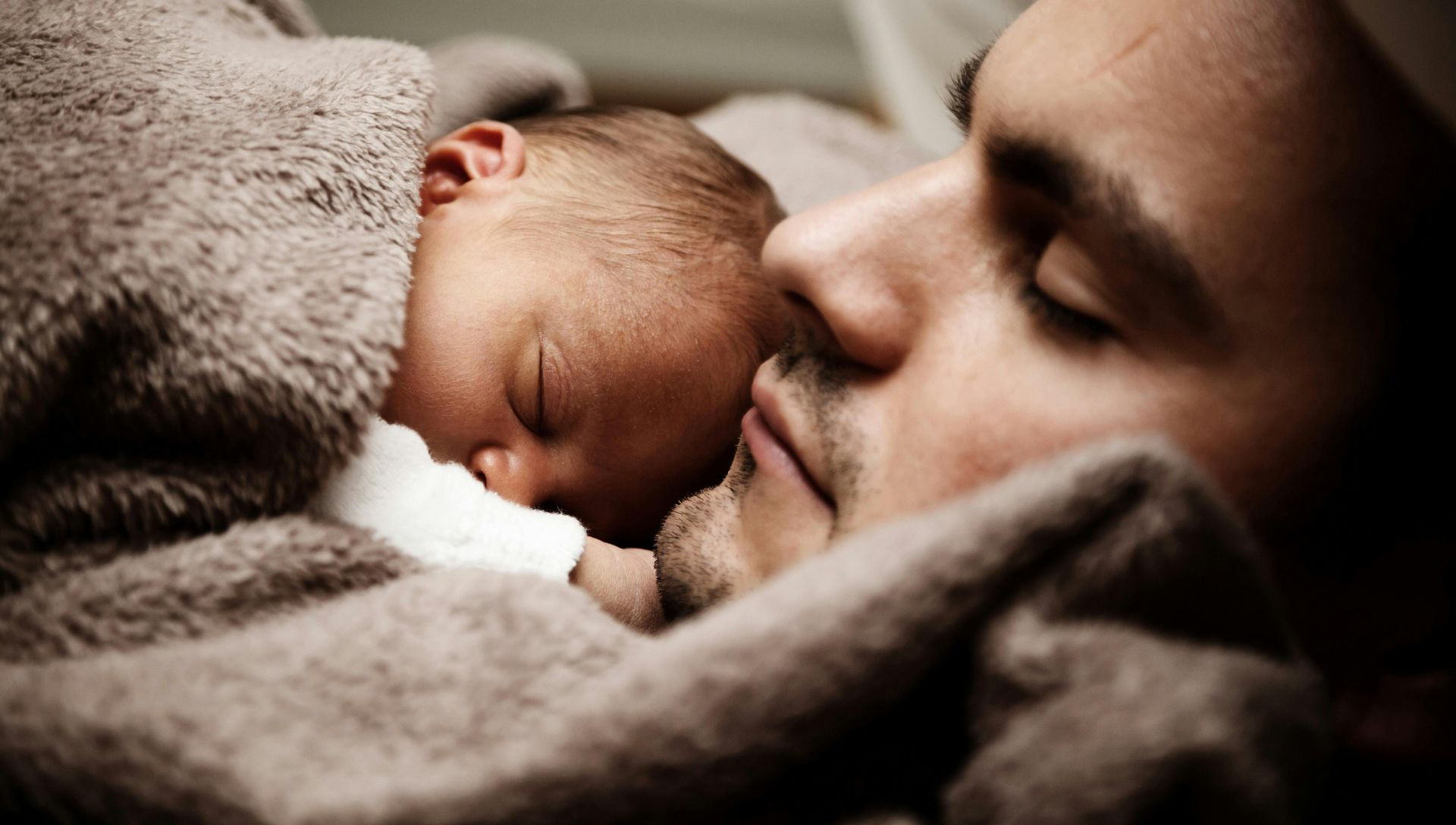 Sleeping baby nestled next to a man; both wrapped in a soft, brown blanket.