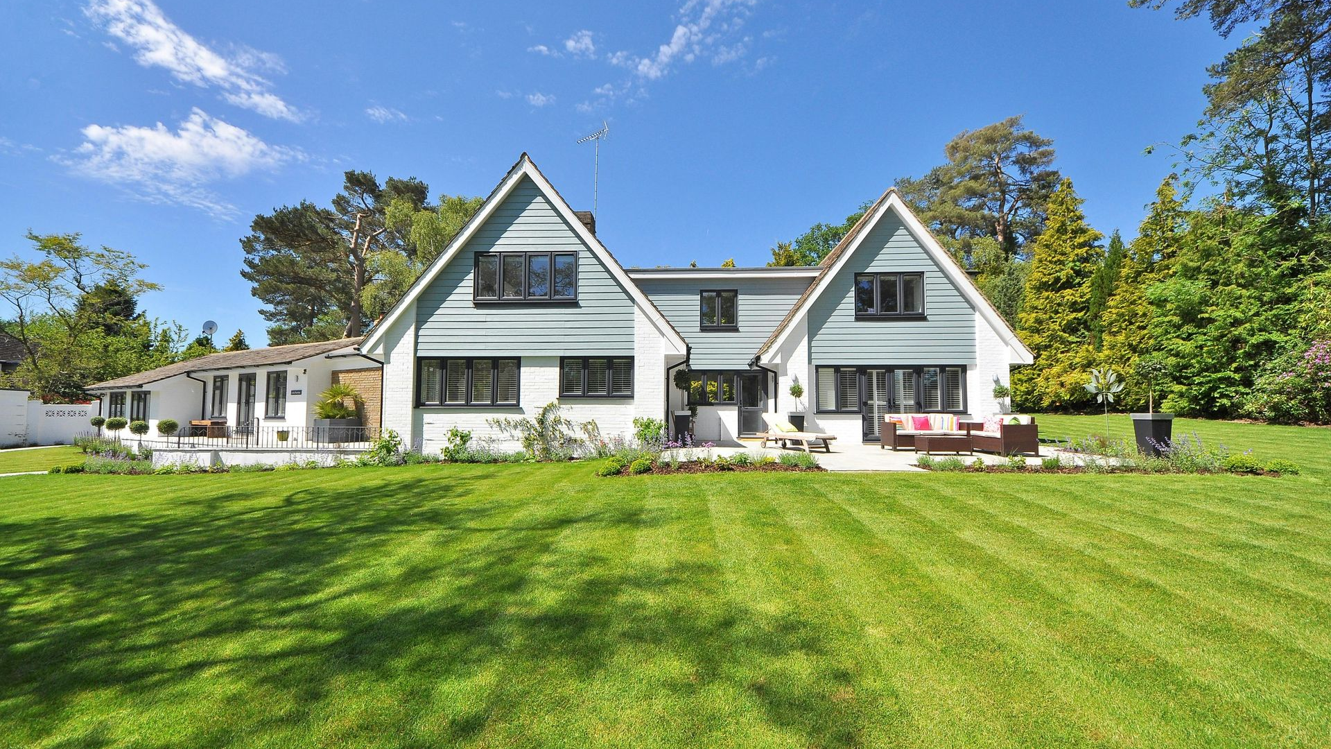 House with white siding and blue gables, sitting on a grassy lawn under a blue sky with trees.