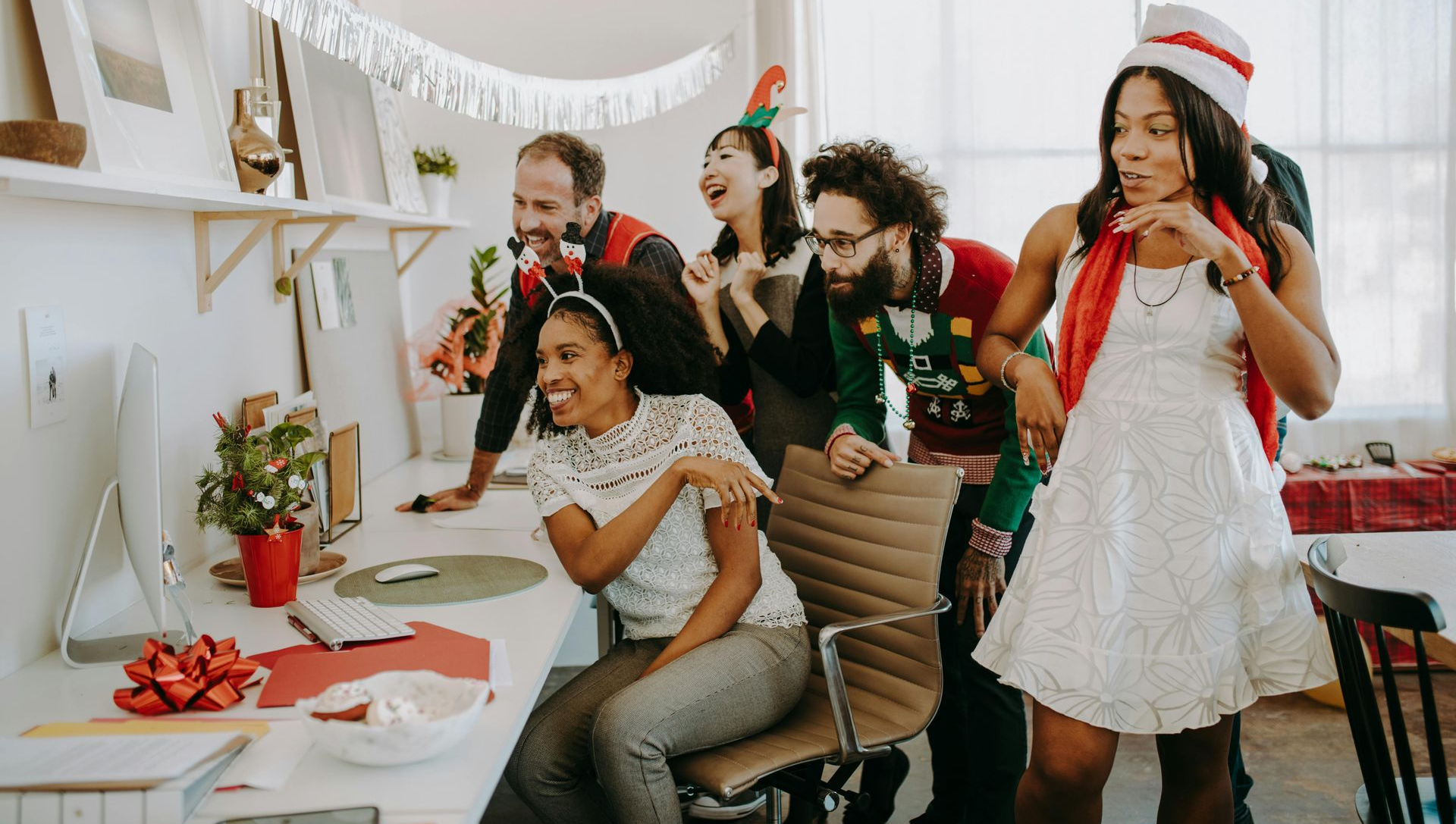 Office workers in festive attire gathered around a computer, laughing and celebrating with Christmas decorations.