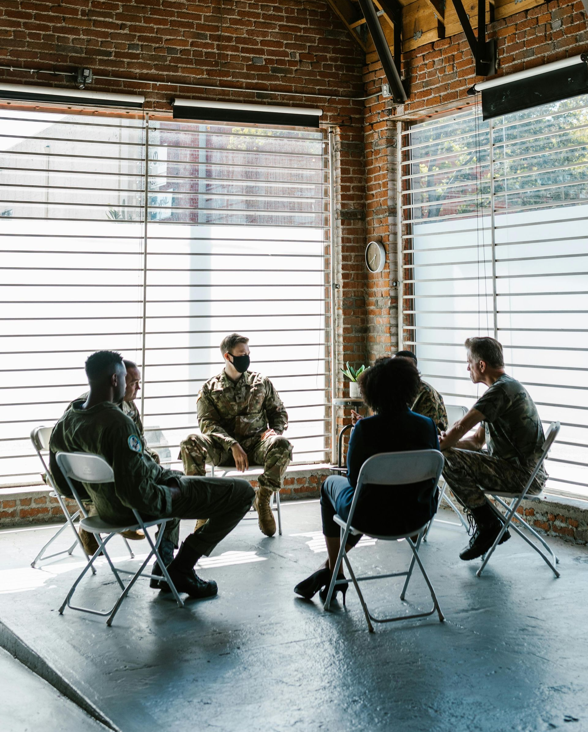 Group of people in a circle, inside a brick building with large windows. Several are wearing camouflage.