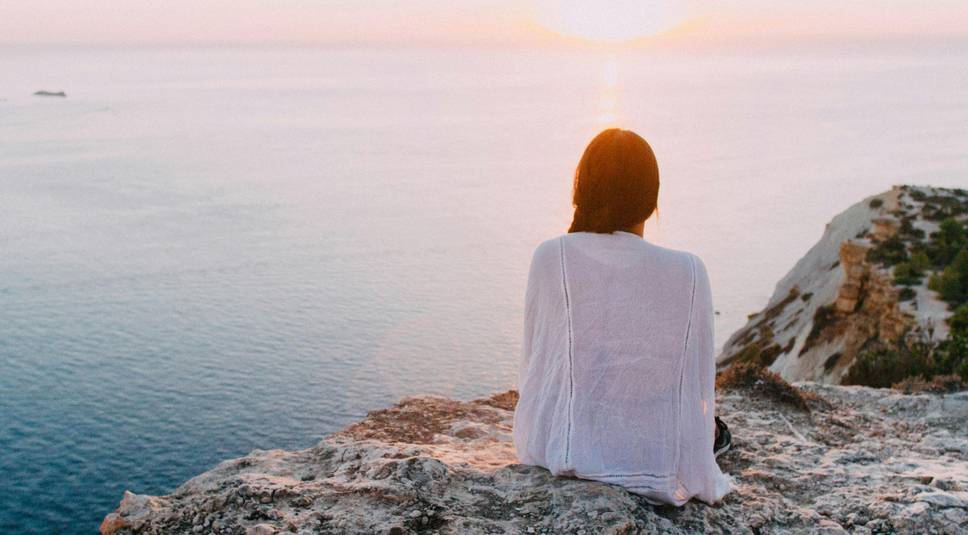 Woman sitting on cliff, facing the sunset over the ocean. Wearing white, serene moment.