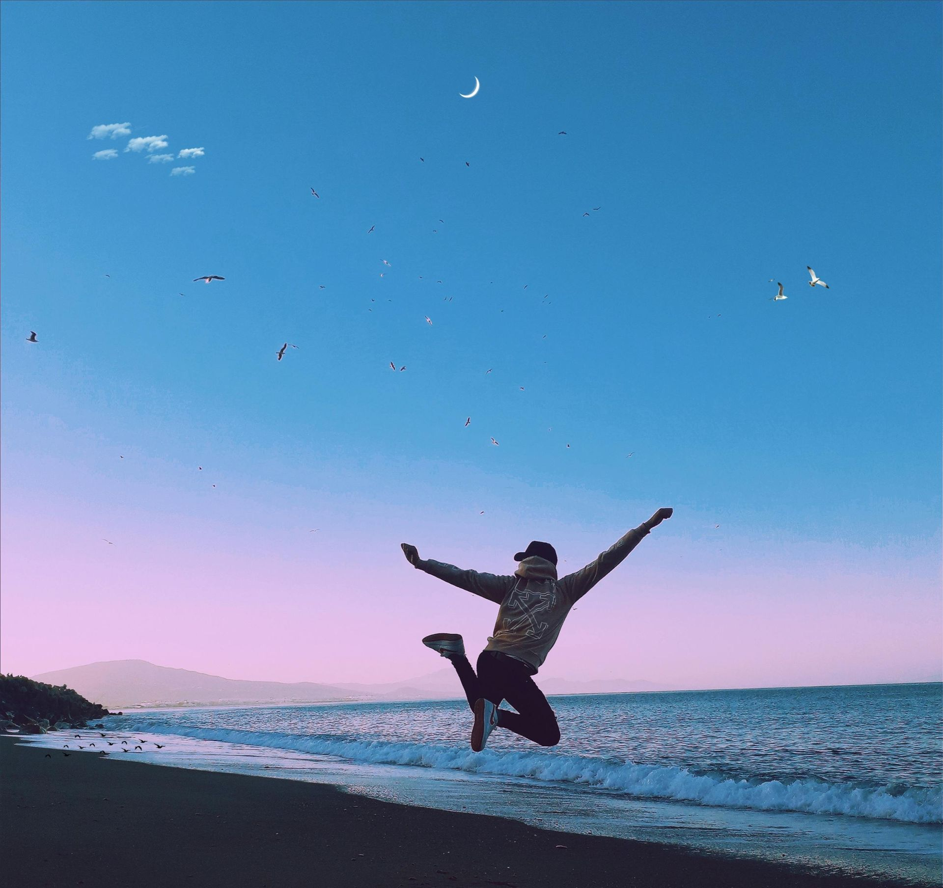 Person jumping on a beach with arms outstretched towards a blue and pink sky with ocean waves.