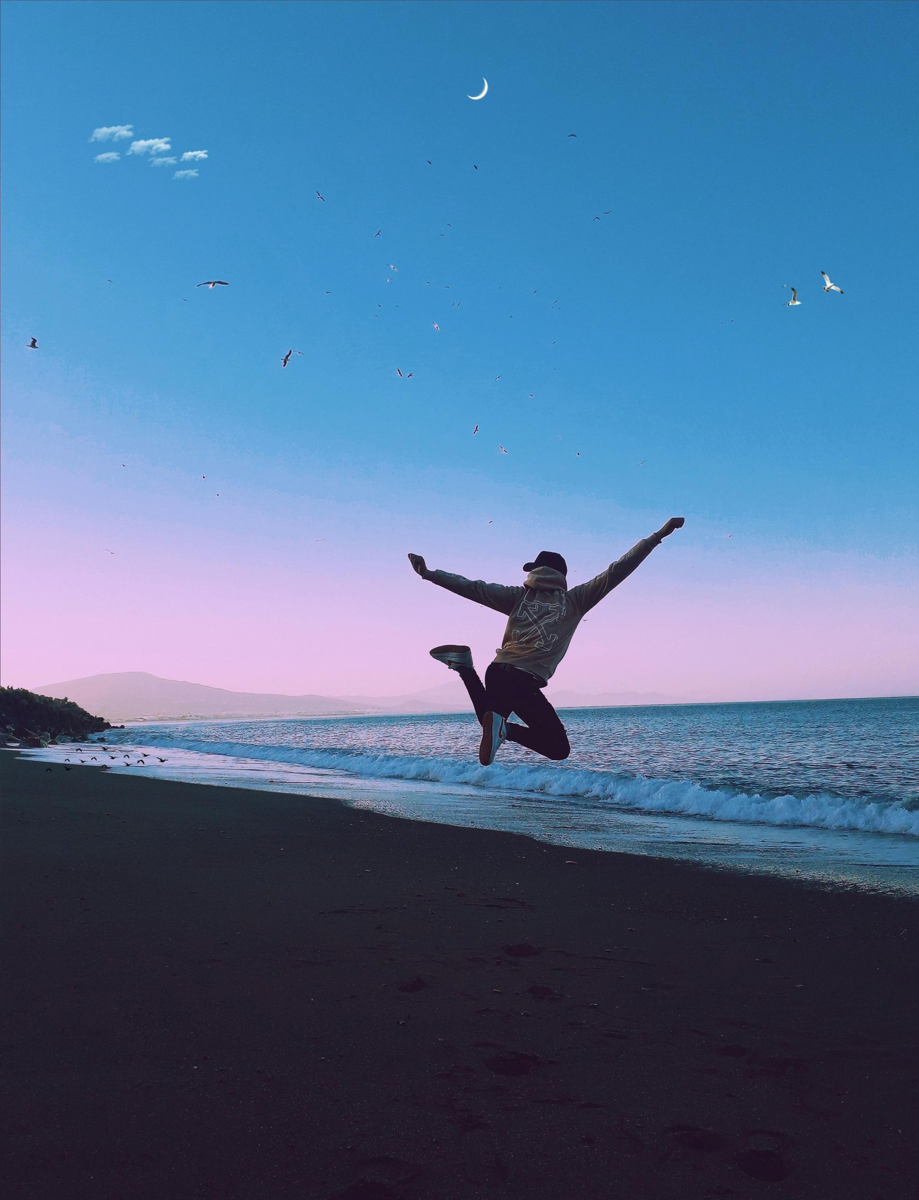 Person jumping on a beach with arms outstretched towards a blue and pink sky with ocean waves.