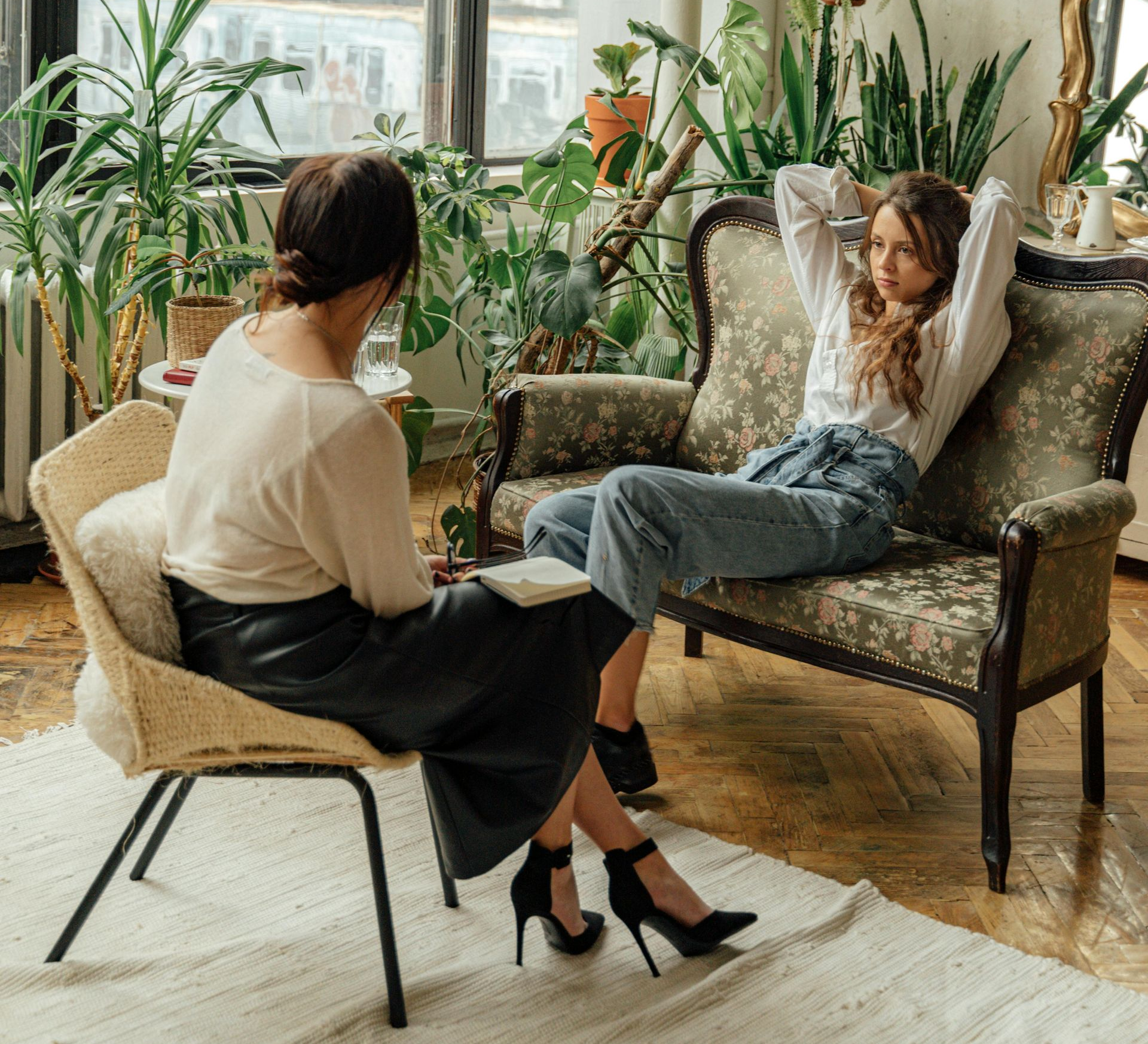 Woman on couch talking to a person seated, possibly a therapist. Room filled with plants.