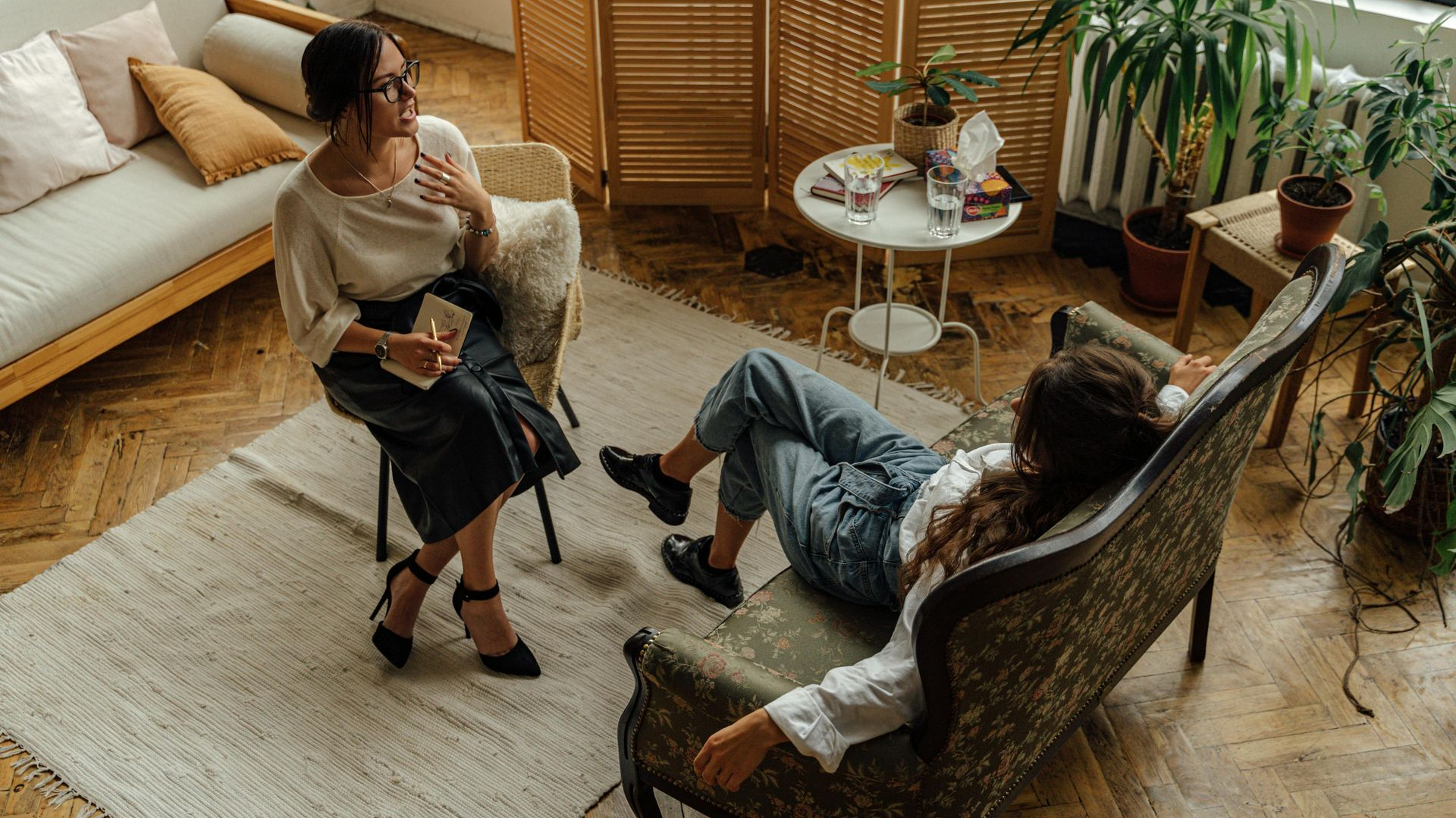 Two women in a therapy session: one seated, talking; the other reclining on a chair, listening. Room with plants and couch.