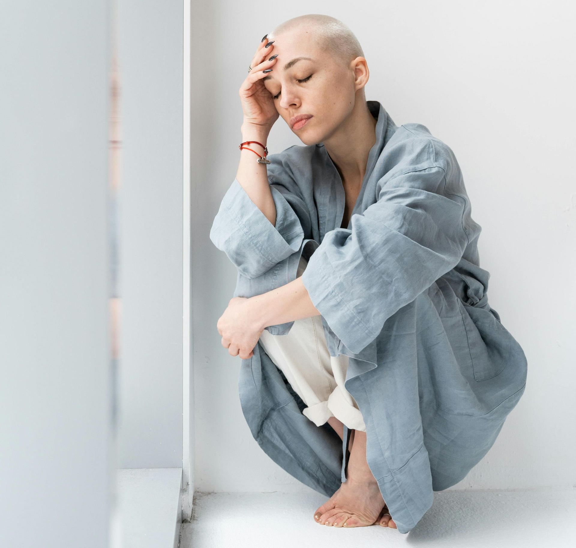 A woman with a shaved head sits hunched, hand on her forehead, by a white wall. She wears a blue robe.