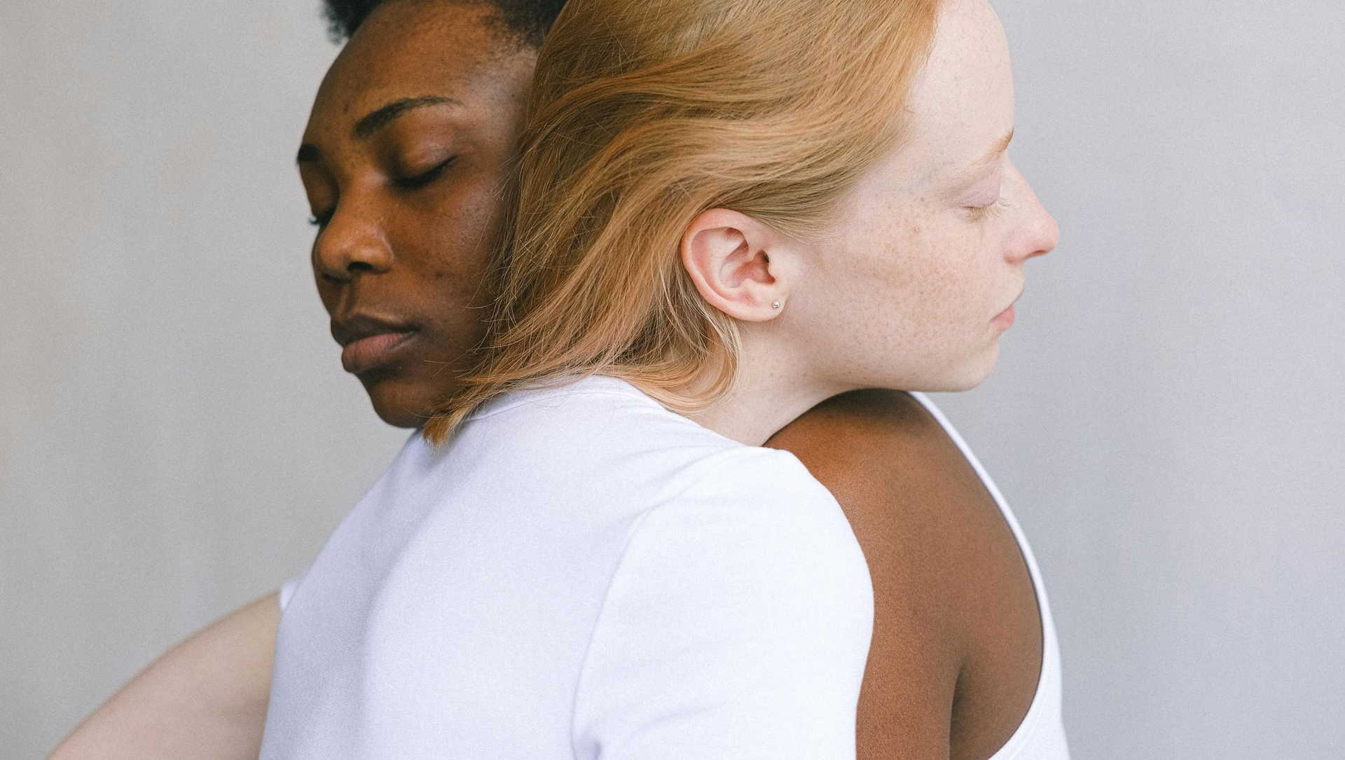 A Black person and a white person embrace, both with eyes closed, wearing white shirts, in front of a gray backdrop.