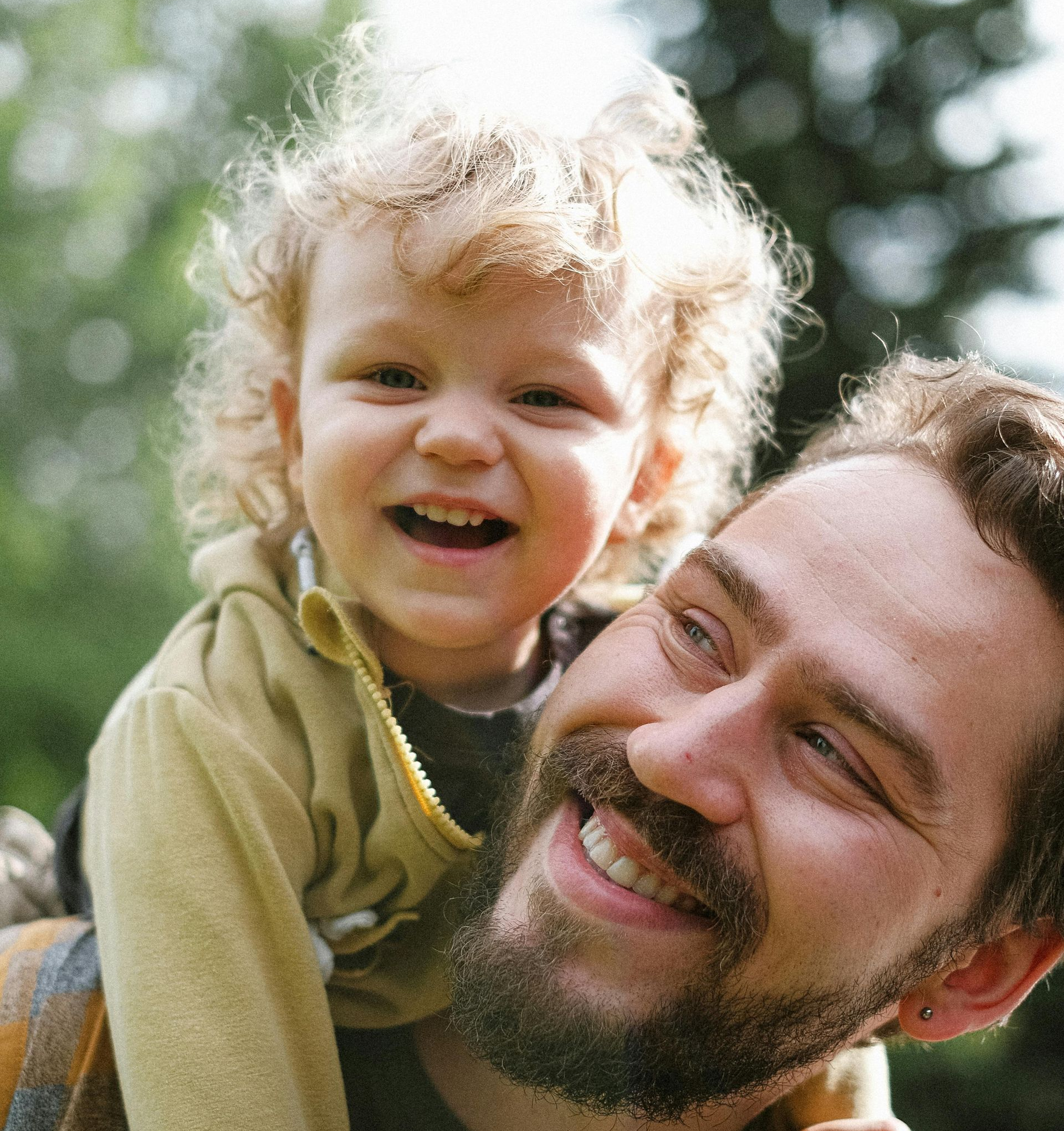 Man with a beard smiling, carrying a smiling child with curly hair on his shoulders, outdoors.