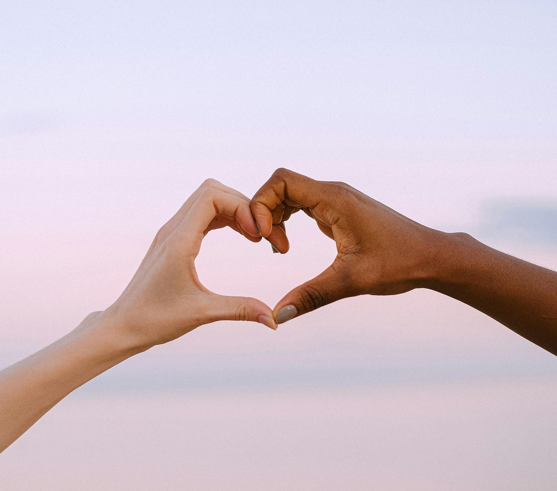 Two hands, one light-skinned and one dark-skinned, forming a heart shape against a pink sky.