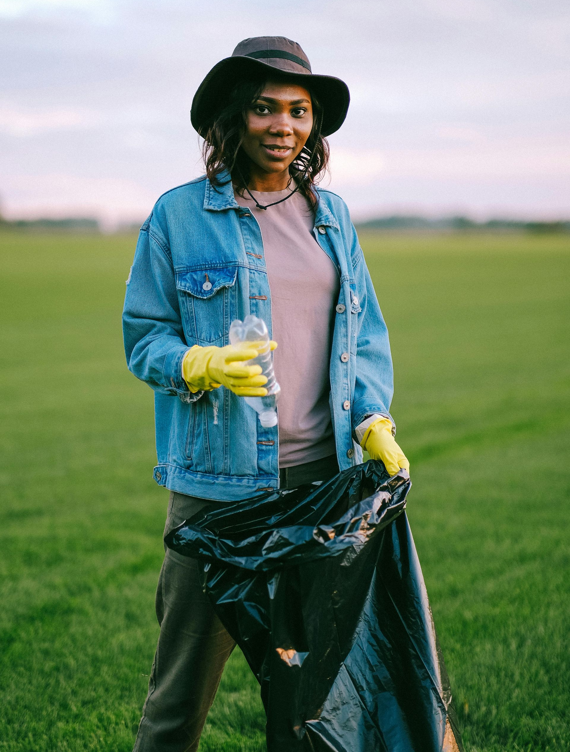 Woman in jean jacket and hat collects trash in a field. Holds a bottle and a trash bag, wearing gloves.