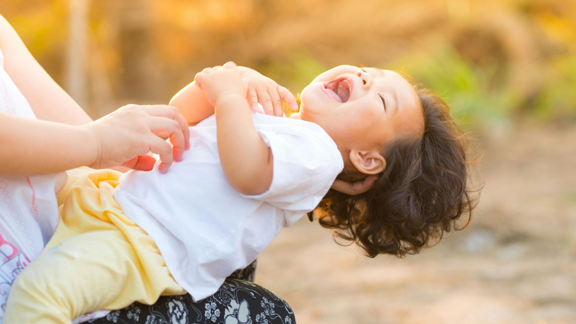 A laughing child is being tickled, outdoors. The child wears a white shirt and yellow pants.