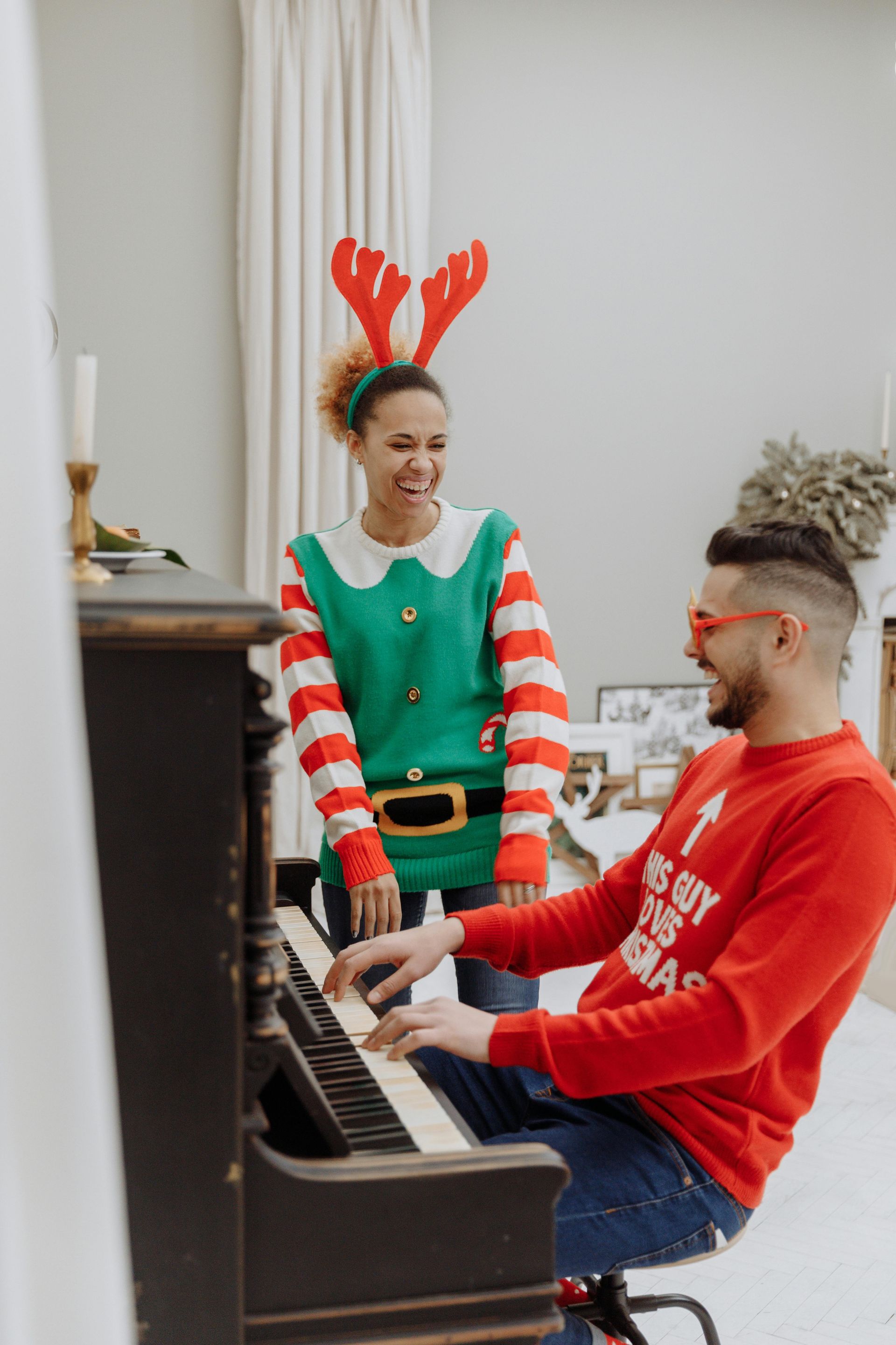 Man plays piano, woman in elf costume laughs. Christmas setting, red and green sweaters.