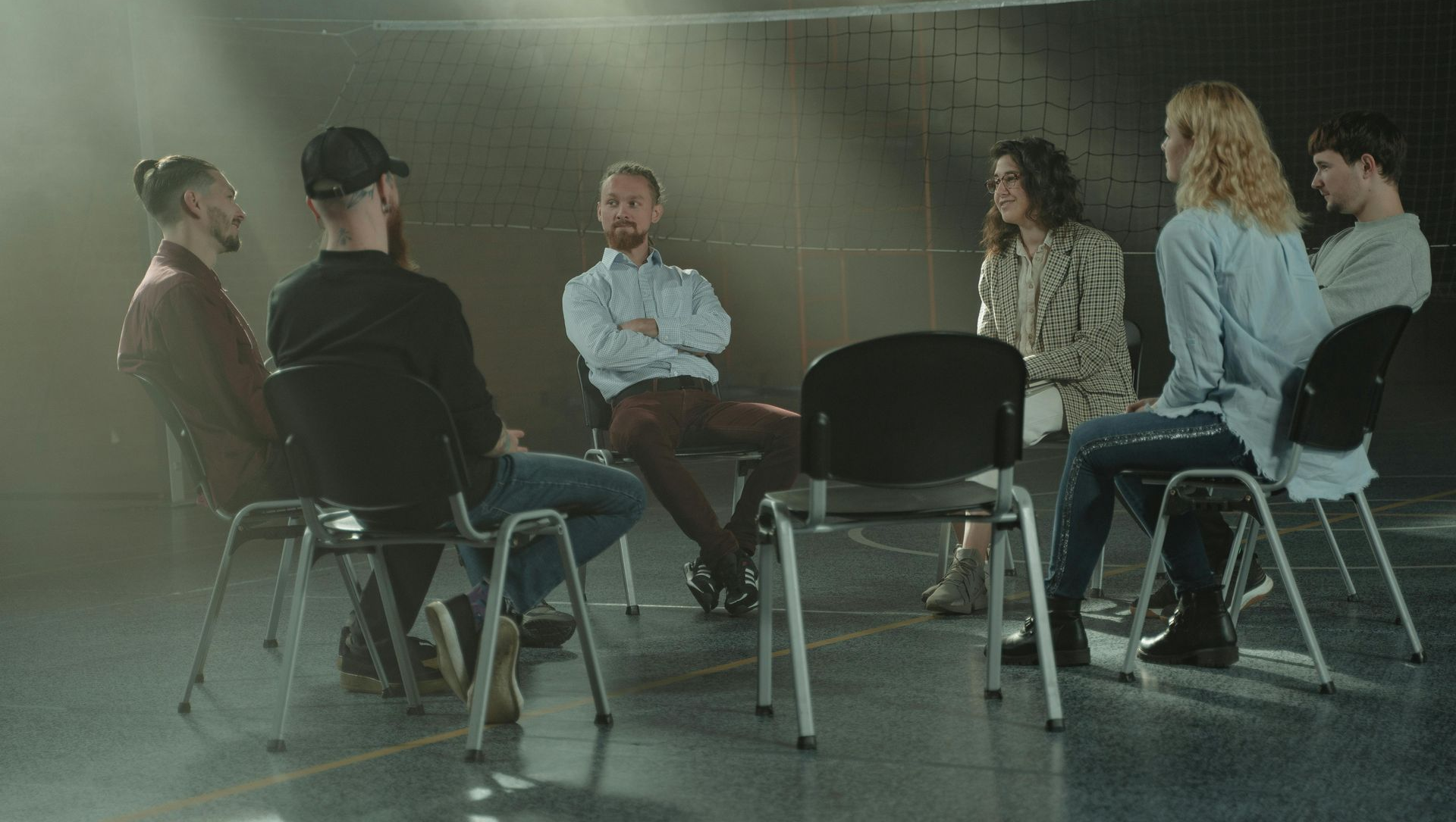 A group therapy session: six people sit in a circle, facing each other and talking in a dimly lit room.