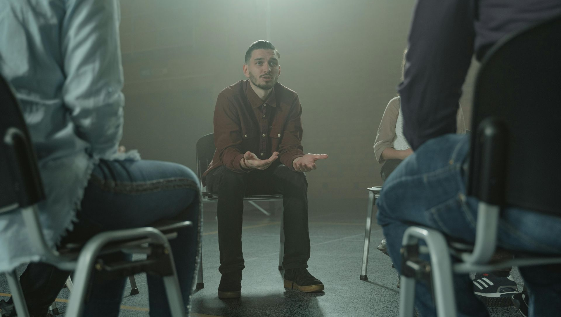 Man in a circle, gesturing as he speaks in a dimly lit setting with other people seated.