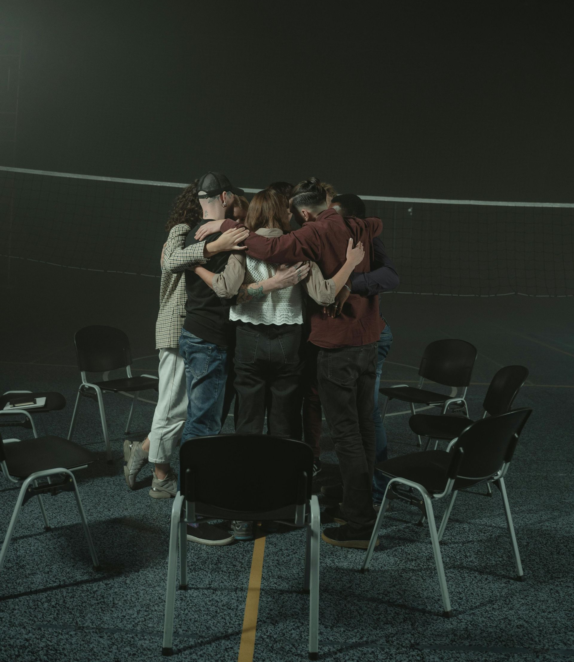 A group of people hugging in a circle in a dark room with chairs.