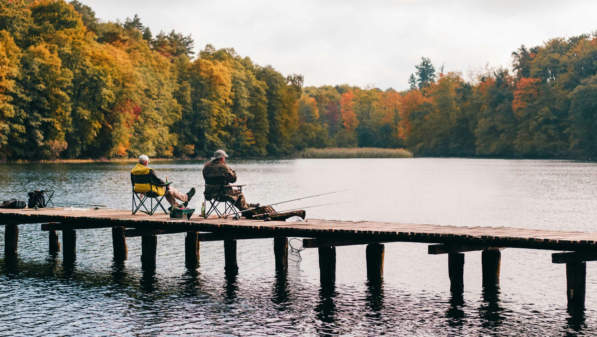 Two people fishing from a wooden pier on a lake surrounded by autumn trees.
