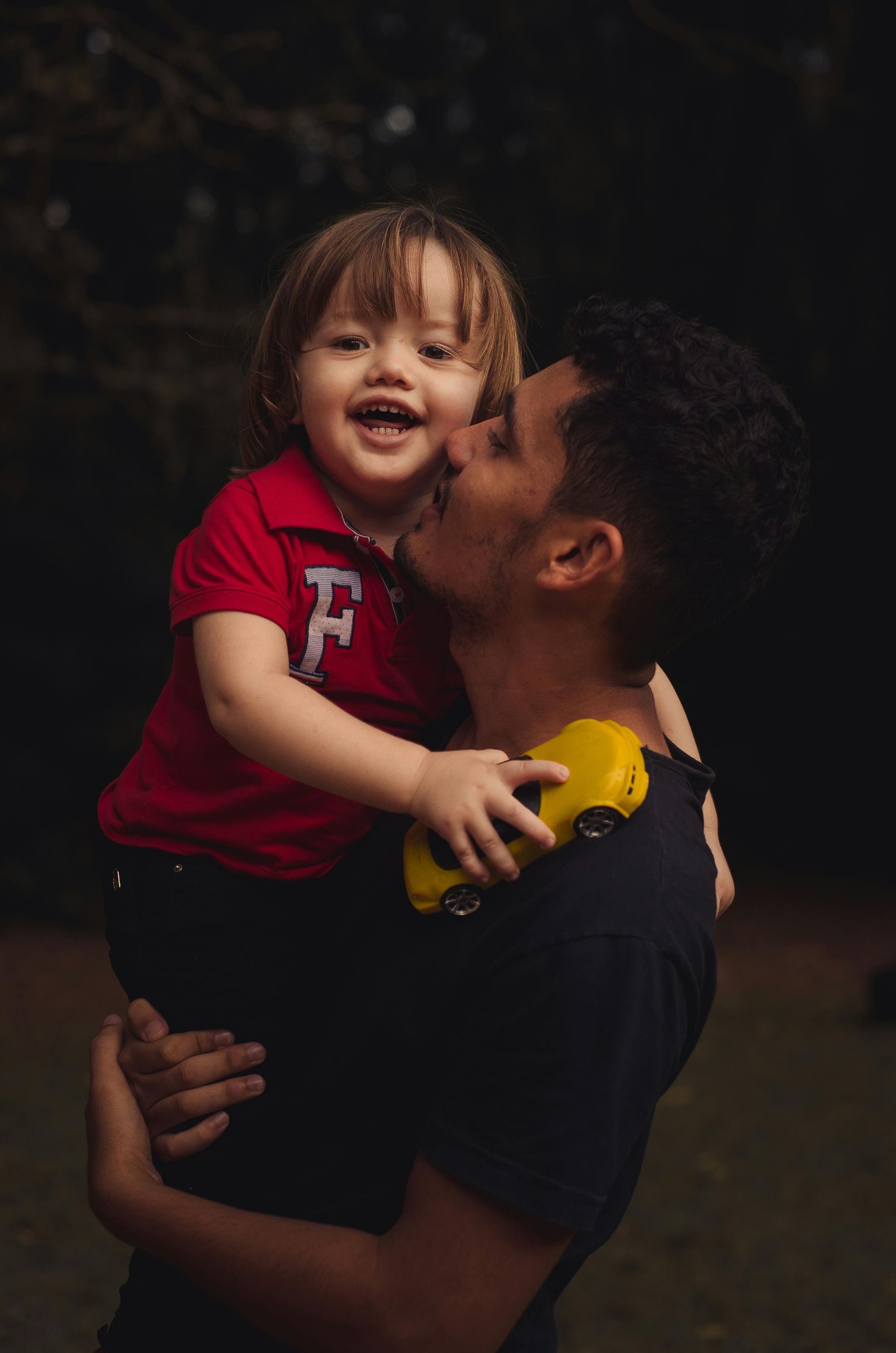 Man holding and kissing a child, both smiling. Child wears a red shirt with a yellow toy car on their shoulder. Dark background.