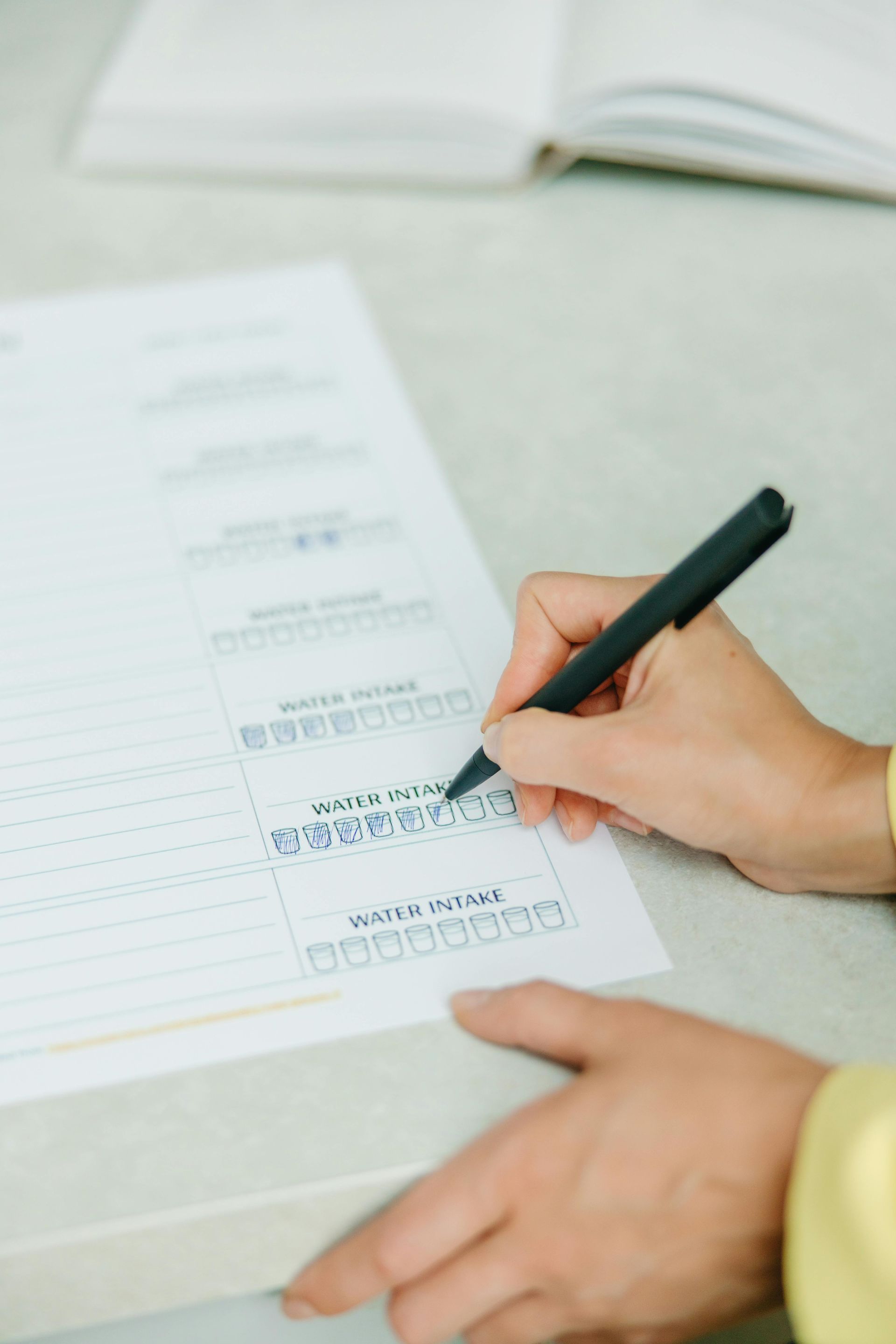 Person filling out a form with a pen. Close-up on hands and paper with check boxes. Light background with open book.
