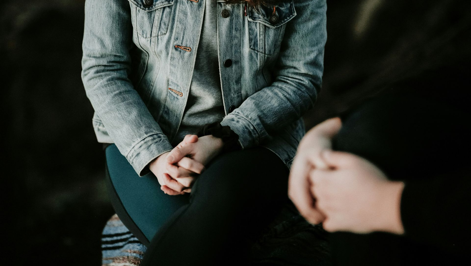 Person in denim jacket with hands clasped, sitting across from another person whose arm is visible.
