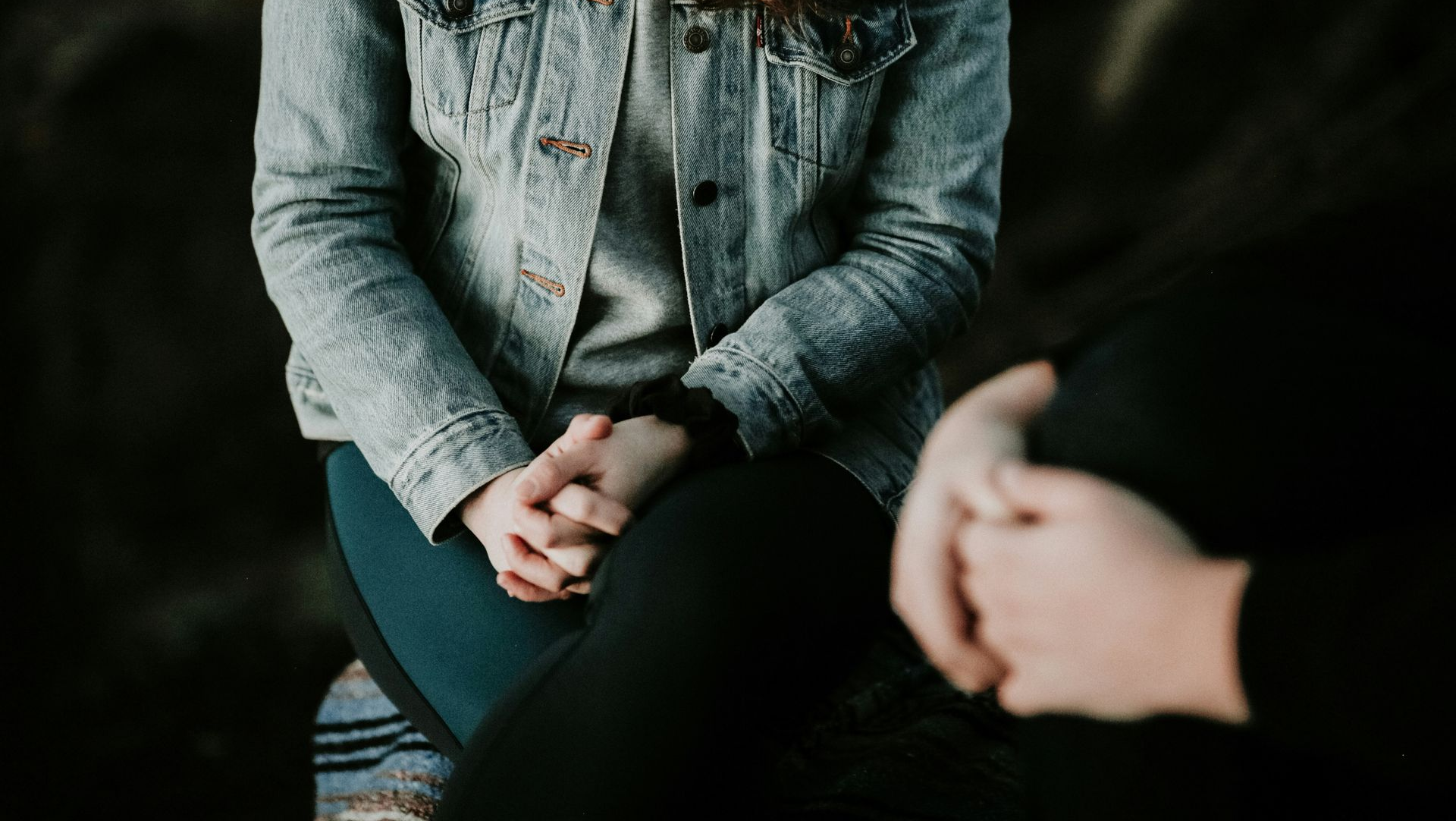 Person in denim jacket with hands clasped, sitting across from another person whose hands are also visible.