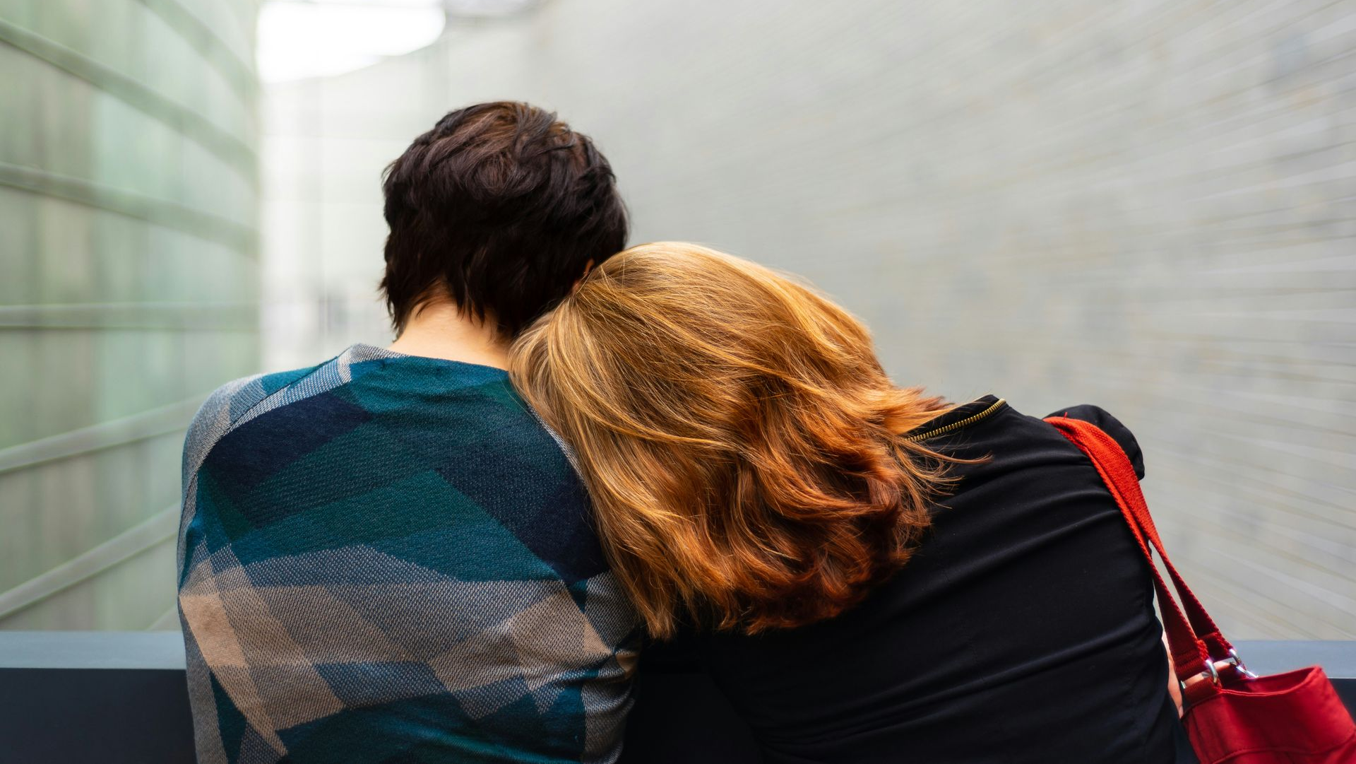 Two people embrace; one rests head on the other's shoulder in modern building.