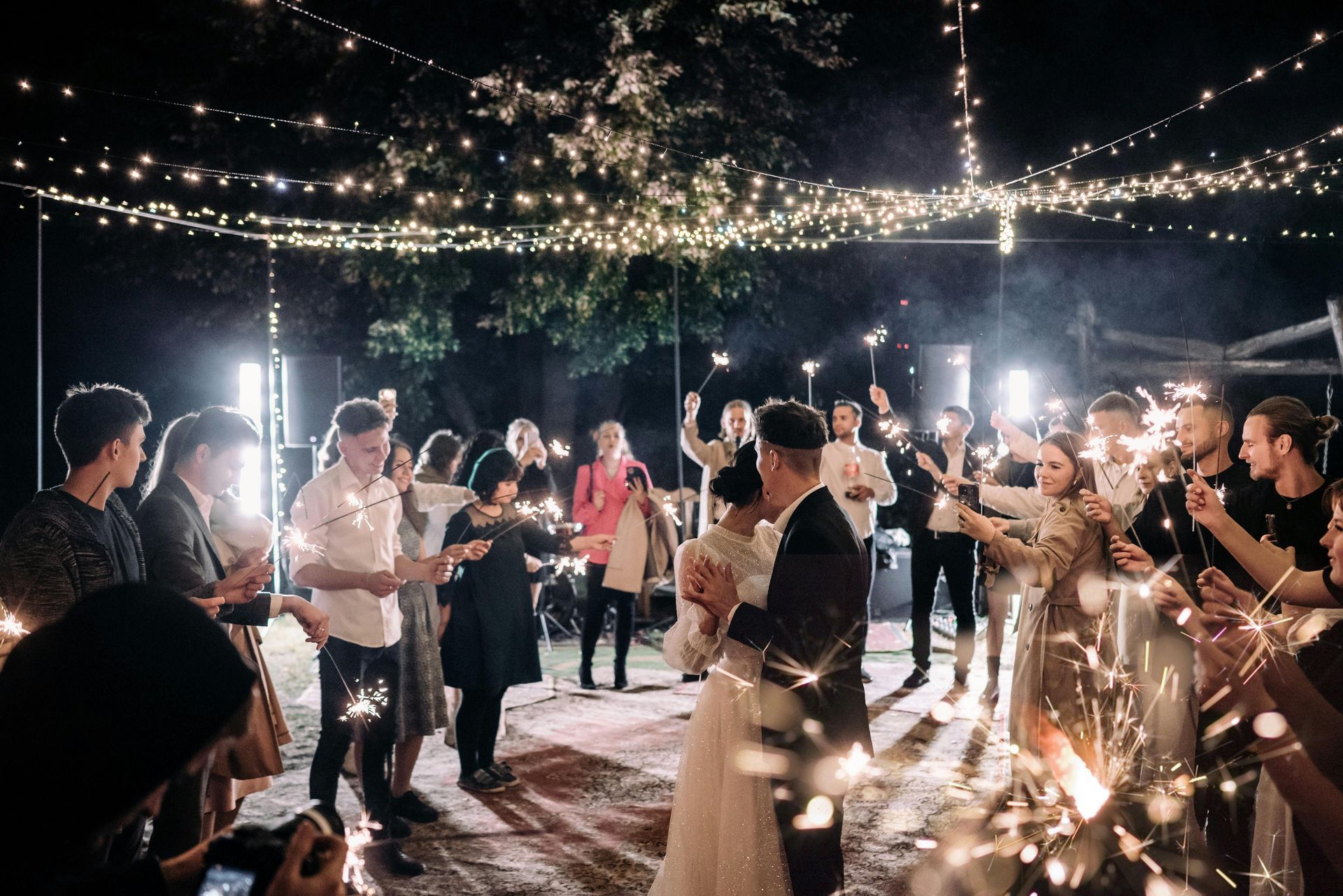 Couple dances beneath string lights as guests wave sparklers at a nighttime wedding.