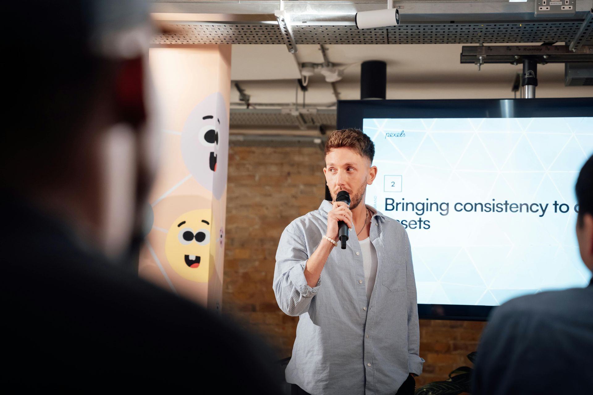 Man speaking into a microphone in front of a screen and audience. Brick wall background.