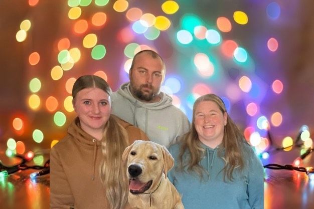 A family is posing for a picture with their dog in front of christmas lights.