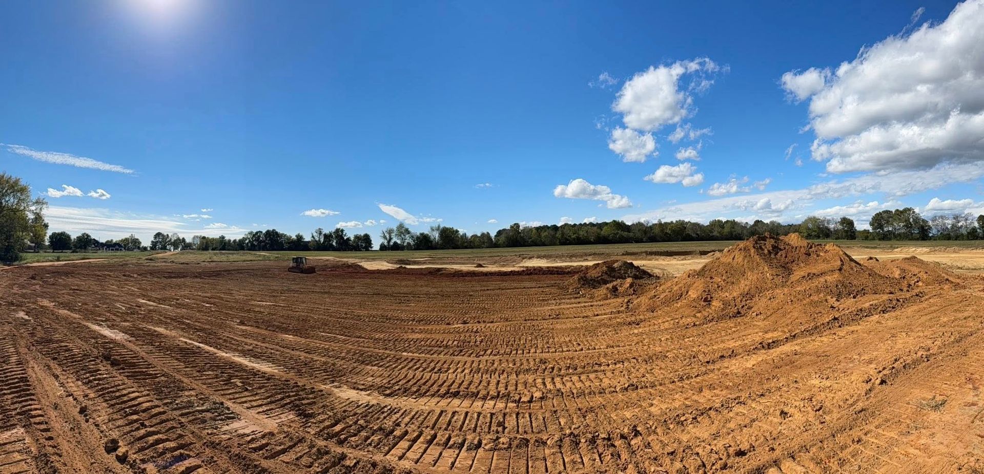 A panoramic view of an excavated construction site featuring piles of brown soil and tire tracks under a sunny blue sky.