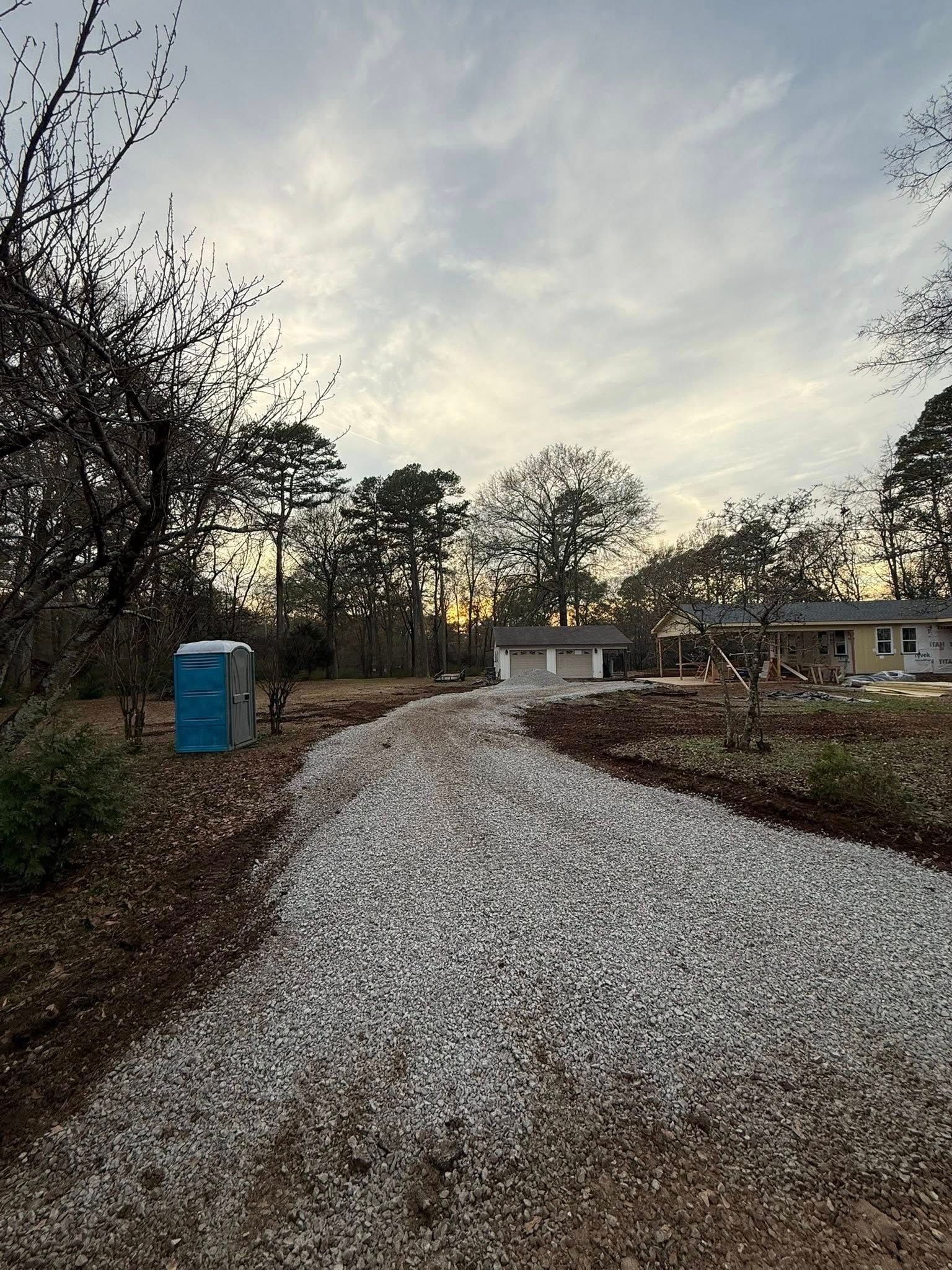 A gravel driveway leads toward a house at sunset, with a portable toilet on the left and surrounding trees.