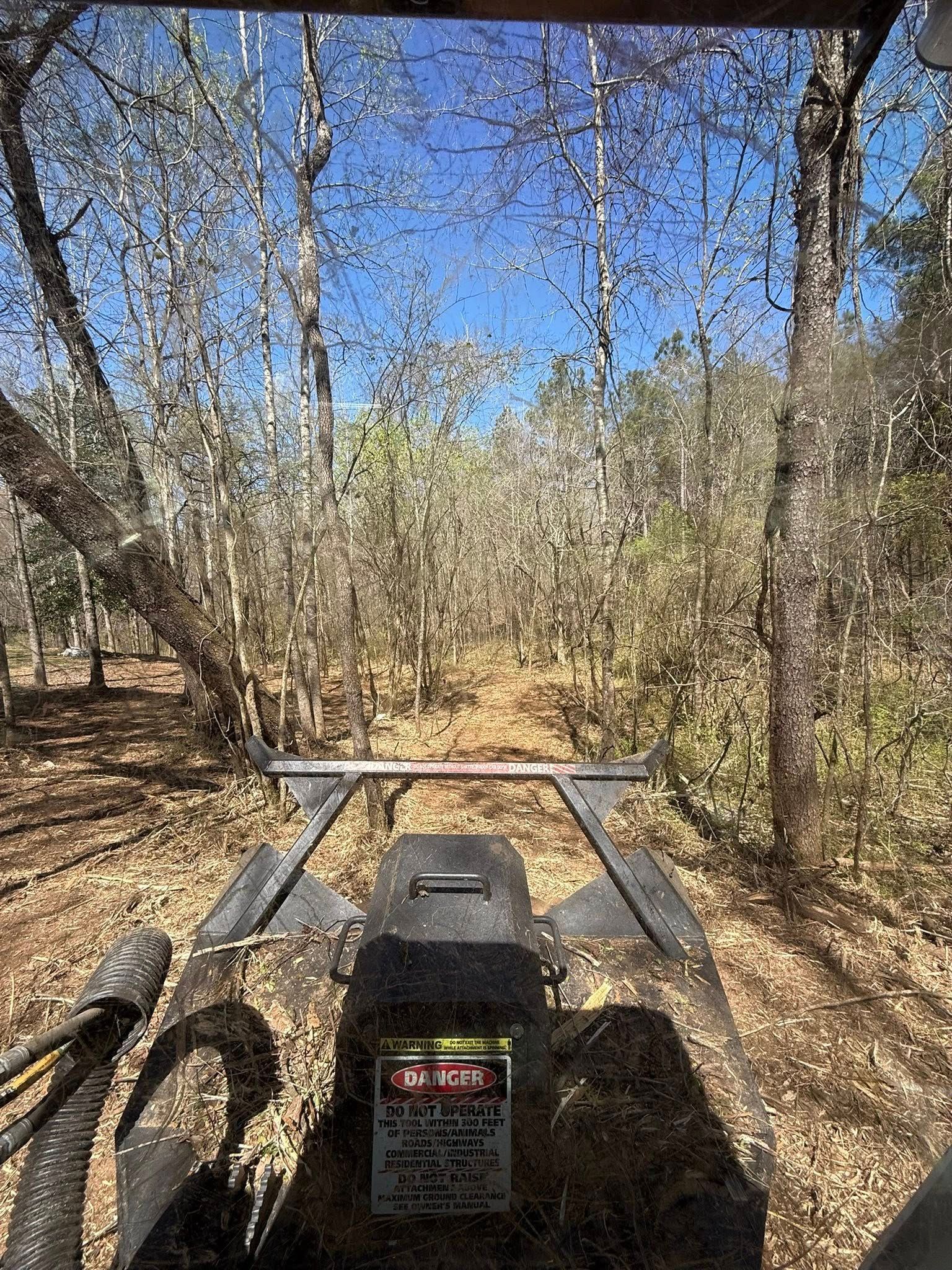 View from the driver's seat of heavy equipment looking down a cleared dirt path through a wooded area under a blue sky.